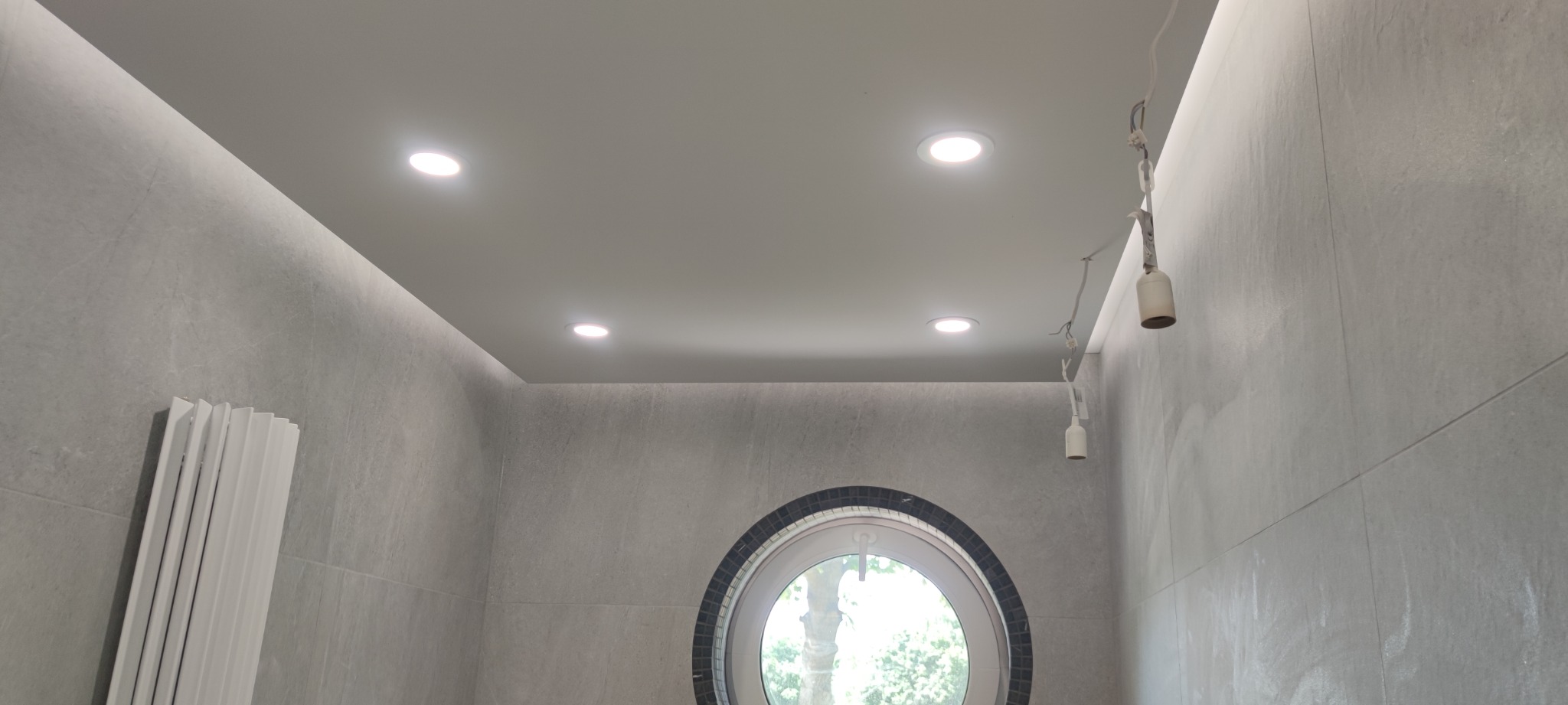 Szara łazienka z okrągłym oknem i oświetleniem sufitowym, widoczne kable od lamp, grzejnik żeberkowy.