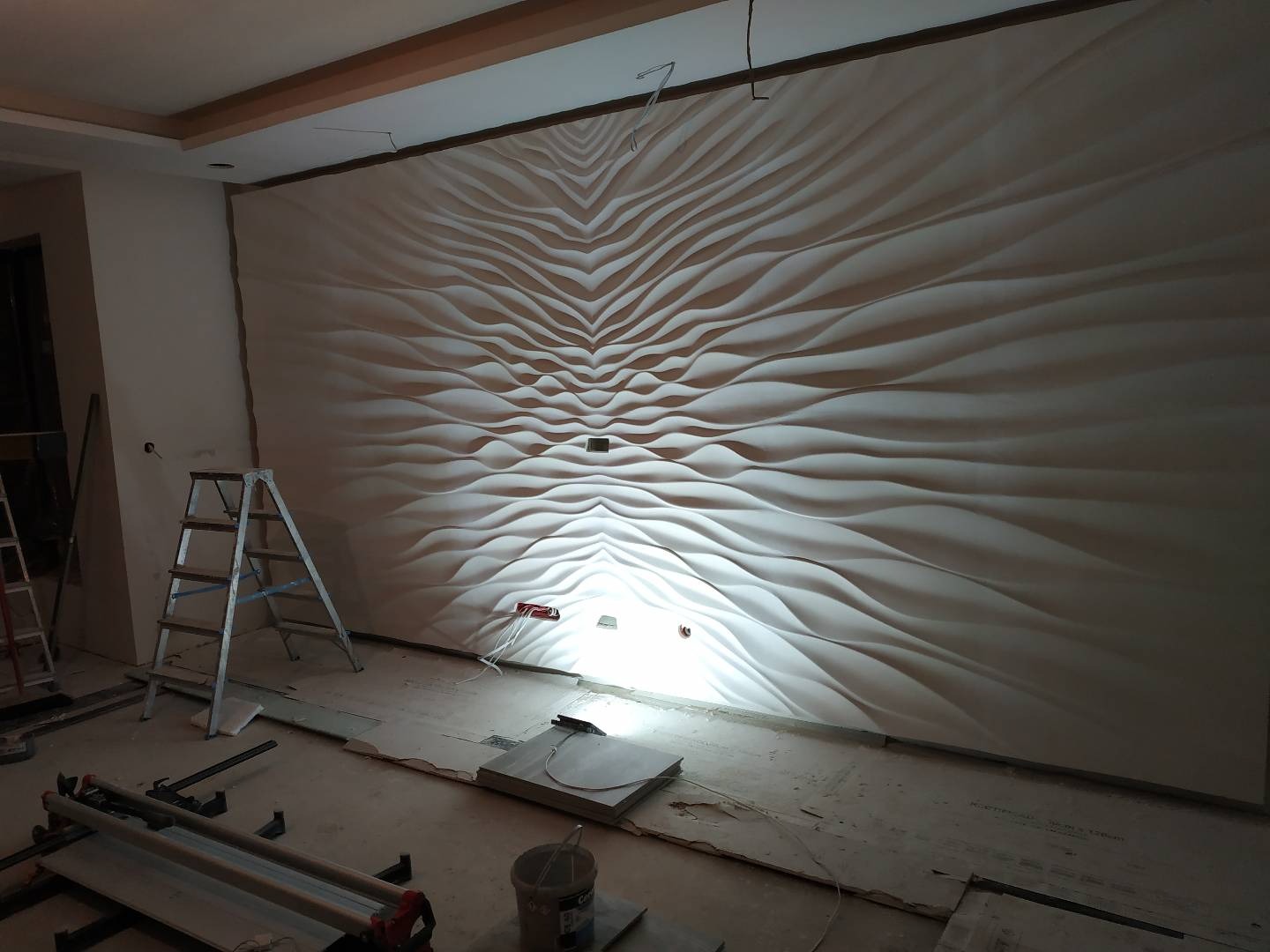 Montaż dekoracyjnej, trójwymiarowej ściany z paneli gipsowych o falistym wzorze w trakcie prac wykończeniowych w mieszkaniu w Warszawie, widoczne narzędzia i drabina.