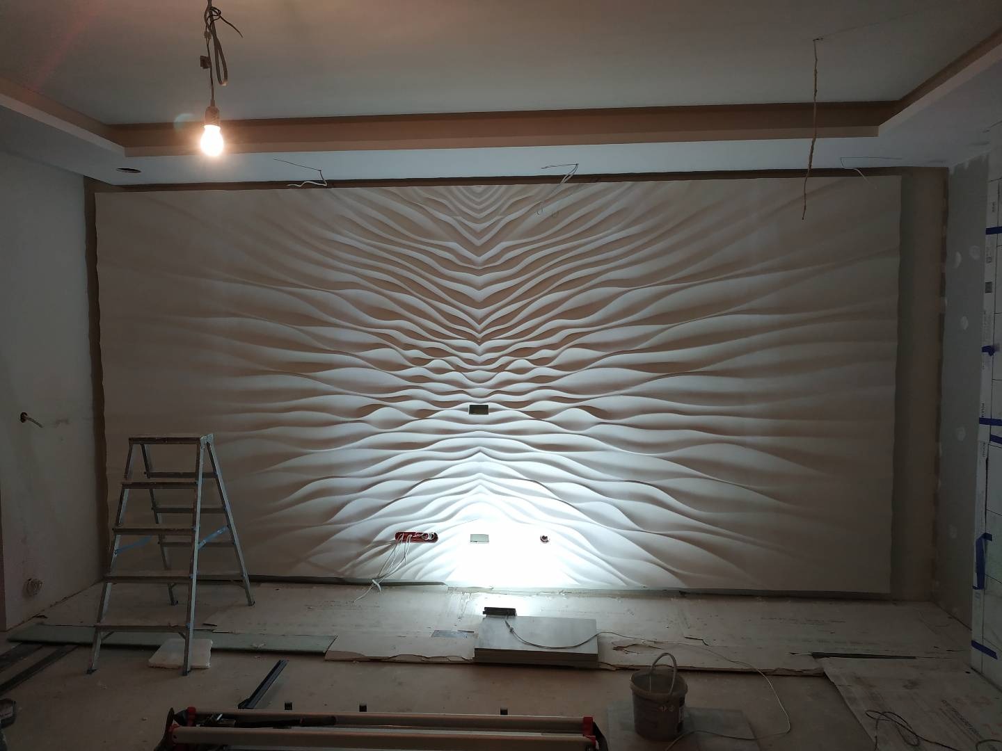 Ściana z dekoracyjnymi panelami 3D w kształcie fal, oświetlona punktowo, w trakcie montażu, widoczna drabina i narzędzia na podłodze.