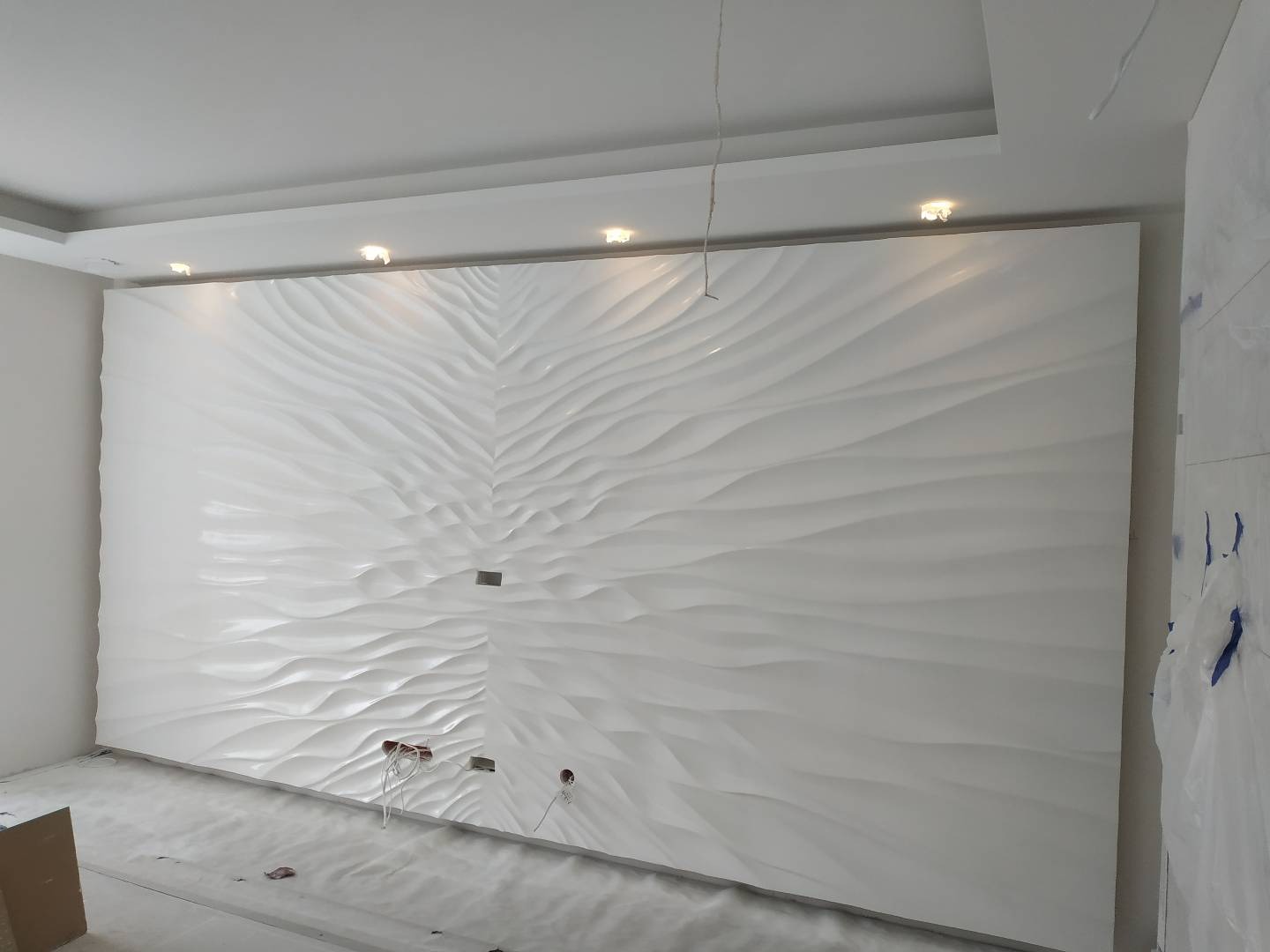Biała ściana dekoracyjna z panelami 3D o falistym wzorze, podświetlona punktowymi lampami sufitowymi, widoczne otwory na gniazdka elektryczne w trakcie montażu.