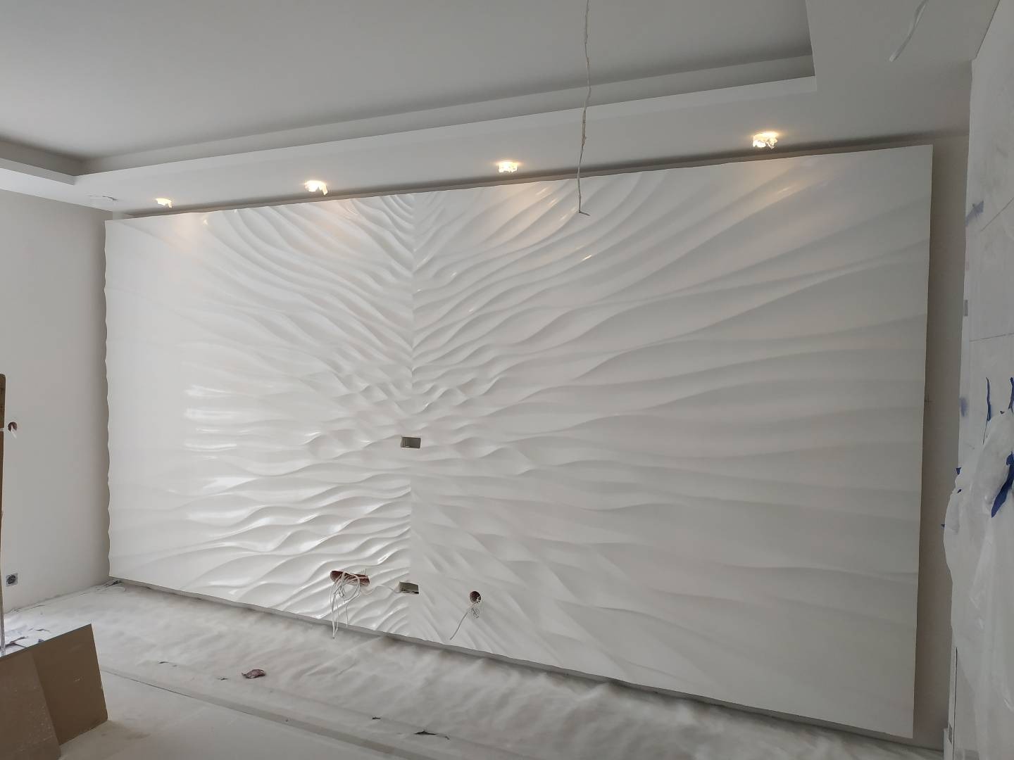 Biała ściana z dekoracyjnym panelem 3D o falistej strukturze, widoczne przygotowane otwory na gniazdka elektryczne i wystające przewody, sufit podwieszany z oświetleniem punktowym.