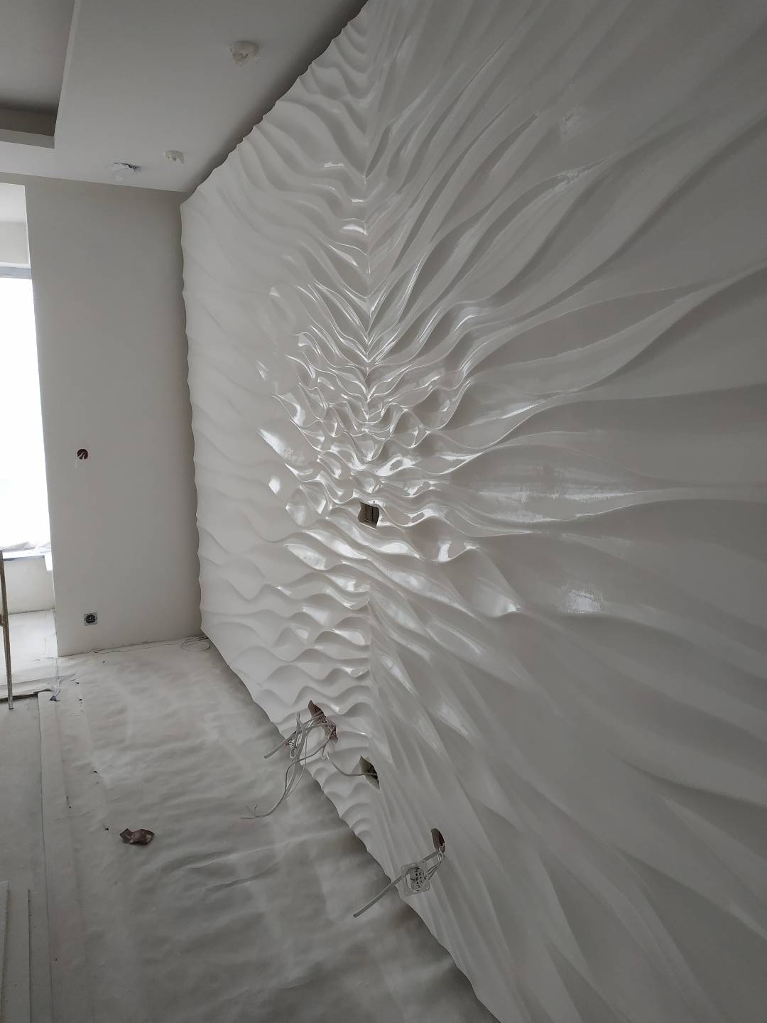 Biała, trójwymiarowa ściana dekoracyjna z falistym wzorem, widoczne wystające kable ze ścian, podłoga zabezpieczona folią malarską.
