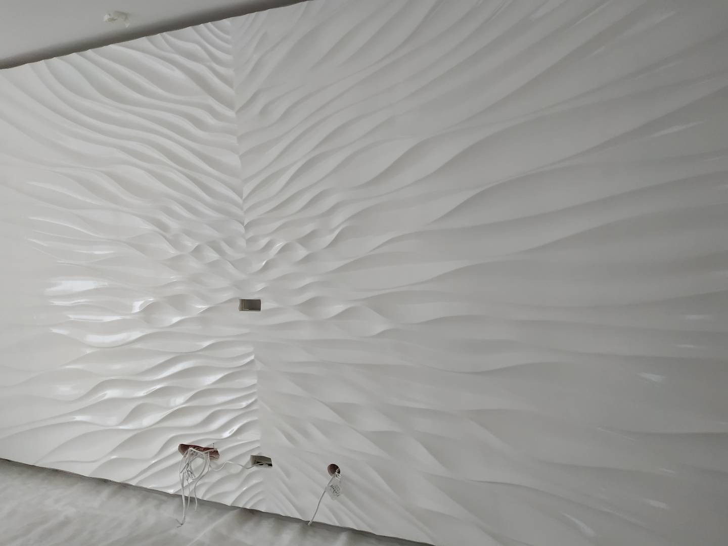 Biała ściana z panelami 3D o falistym wzorze, widoczne otwory pod gniazdka elektryczne z wystającymi przewodami, surowy stan przed montażem osprzętu.