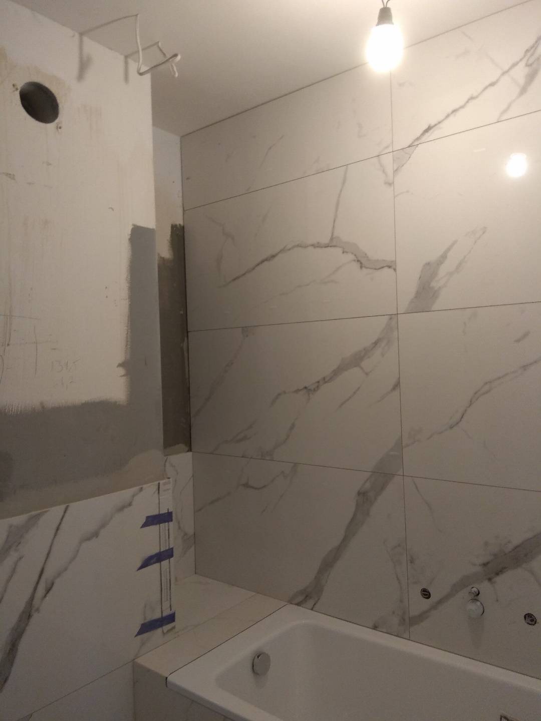 Białe płytki imitujące marmur na ścianie łazienki w trakcie montażu, z widocznymi fragmentami surowej ściany i niebieską taśmą maskującą