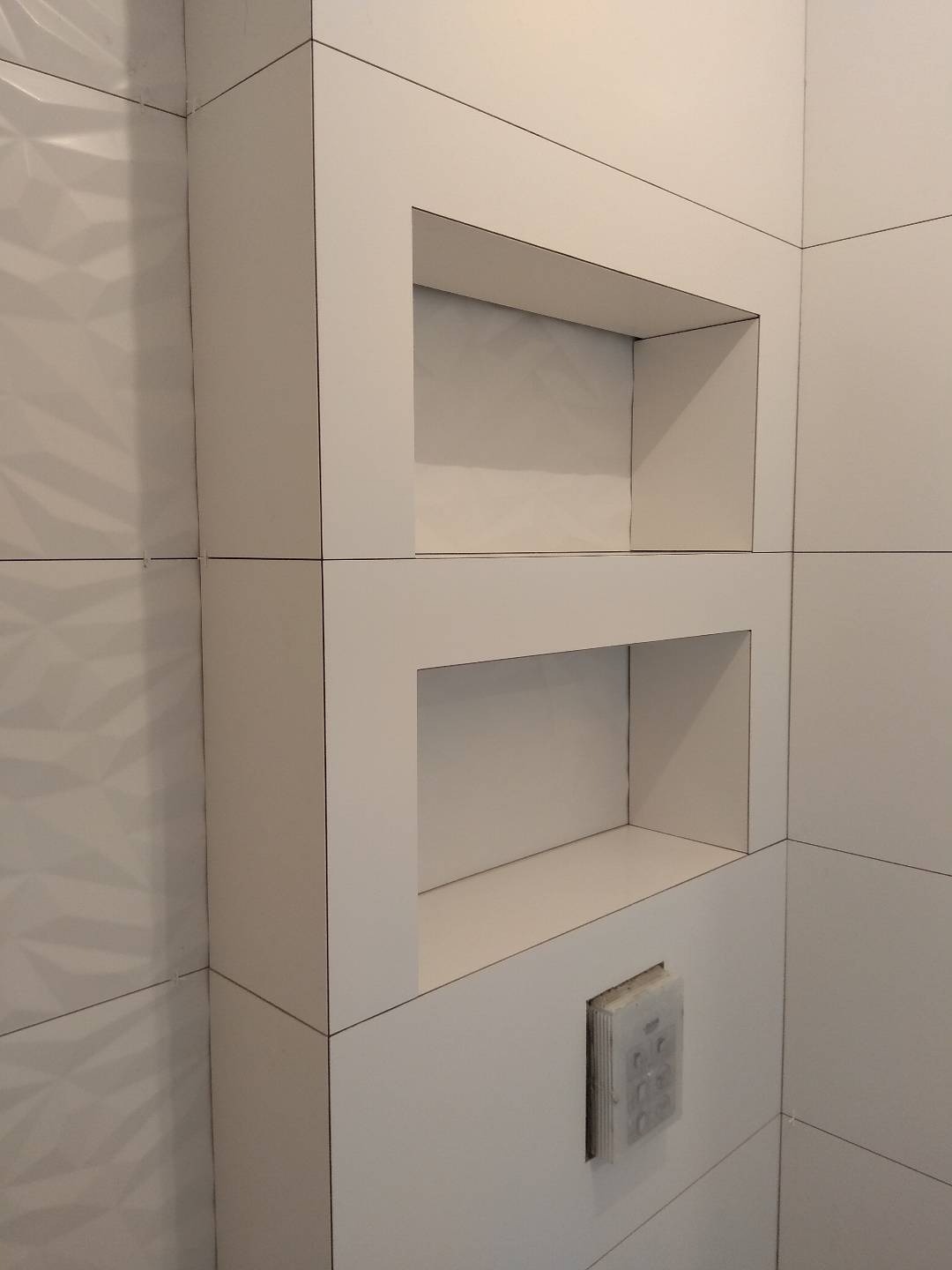 Biała łazienka z wnękami w ścianie wyłożonej płytkami, widoczny przycisk spłuczki pod dolną wnęką, wzór 3D na płytkach po lewej stronie.