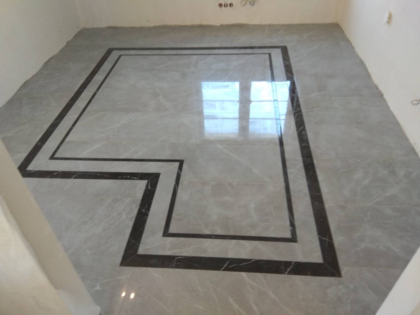 Ułożona podłoga z szarych płytek imitujących marmur, z dekoracyjnym obramowaniem z ciemniejszych płytek, odbicie okna w powierzchni podłogi.