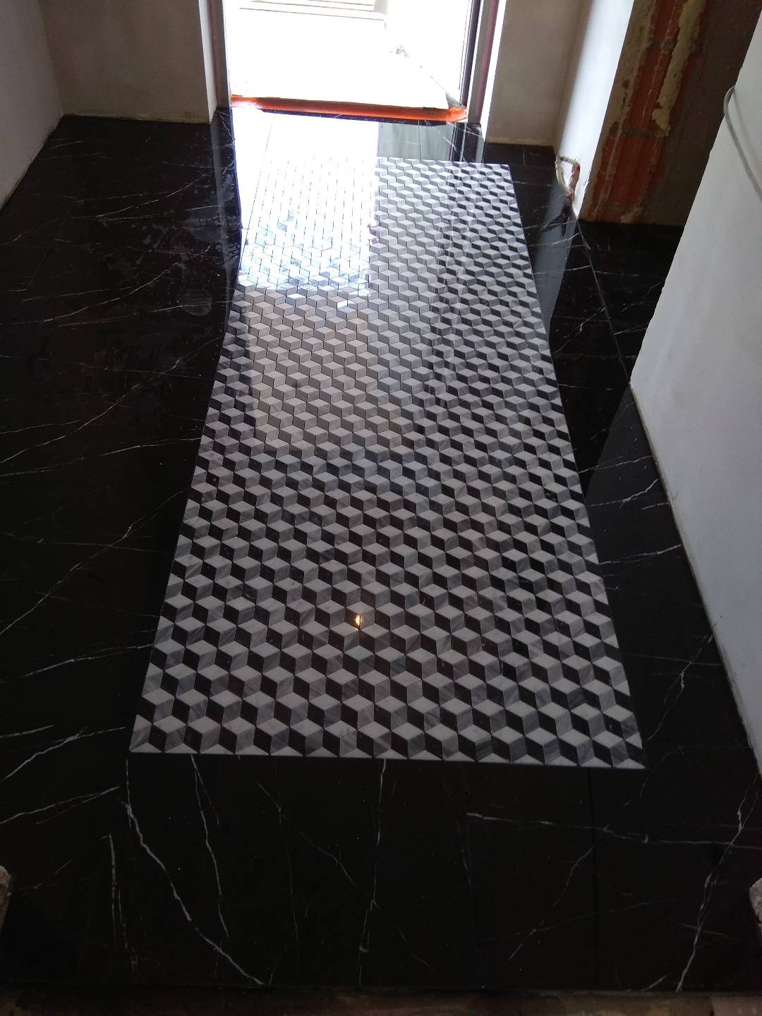 Ułożona podłoga z czarnego marmuru z białymi żyłami, z wstawką z płytek w geometryczny wzór 3D w odcieniach szarości, prowadząca do wejścia.