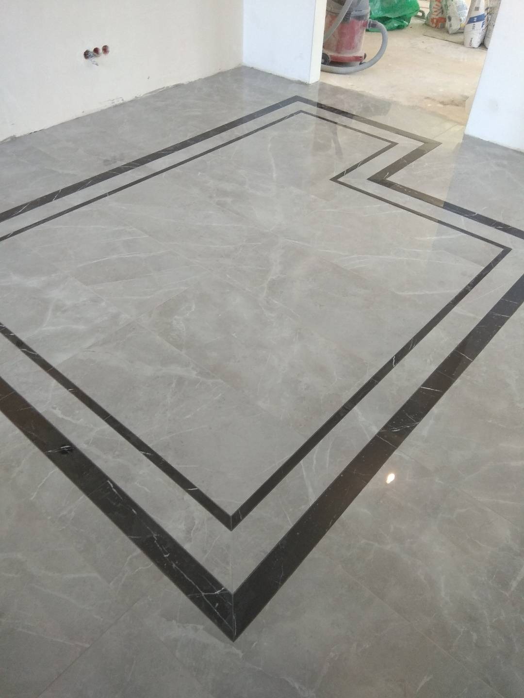 Ułożona podłoga z szarych płytek imitujących marmur z dekoracyjnym obramowaniem z ciemniejszych płytek, widoczny odkurzacz przemysłowy w tle.