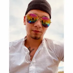 Mężczyzna w czapce i okularach z tęczowymi odblaskami, na których widoczny jest odbity krajobraz. Na koszuli widoczny tatuaż ptaka.