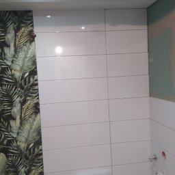 Remonty - Łazienka w trakcie wykańczania: białe, błyszczące płytki ułożone poziomo na ścianie, fragment ściany z tapetą w tropikalne liście, widoczne niedokończone przyłącza wodne i materiały budowlane...