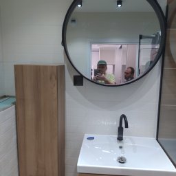 Remonty - Ukończona łazienka z umywalką na szafce z drewnianym frontem, wiszącą szafką obok i okrągłym lustrem, w którym odbijają się dwie osoby.