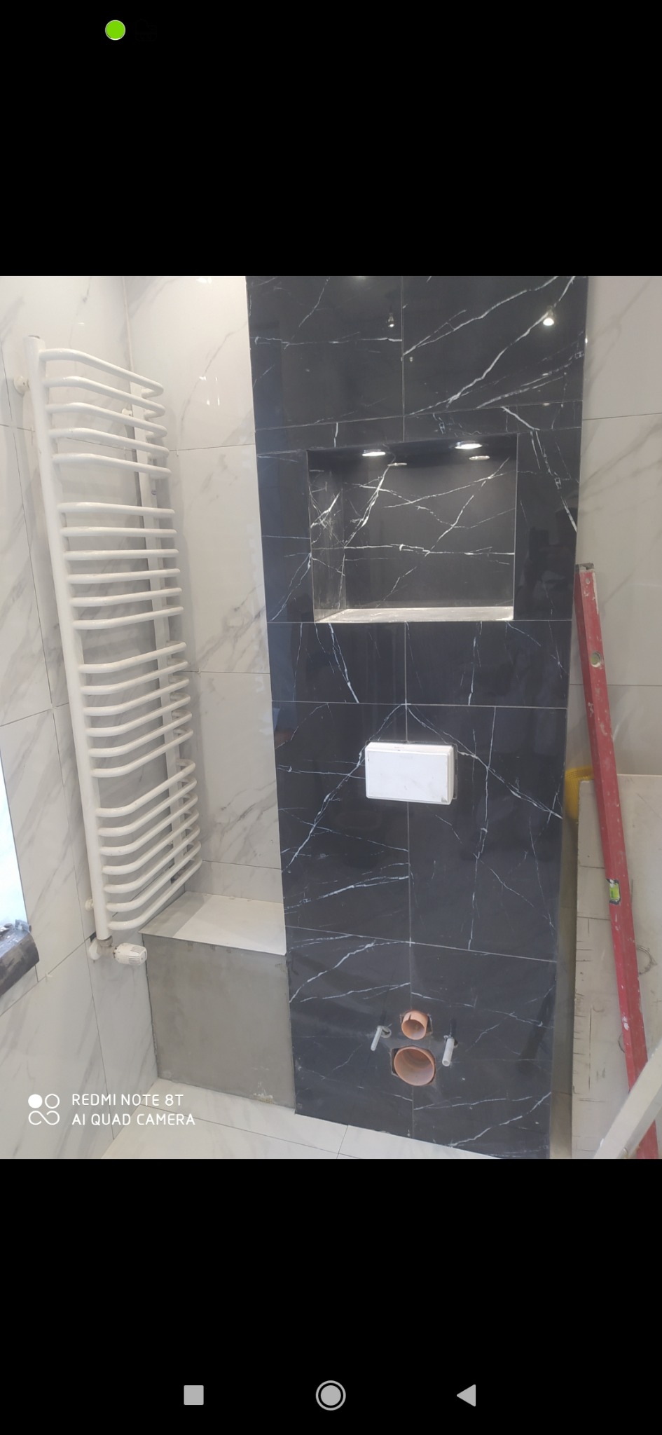 Częściowo wykończona łazienka z czarną marmurową ścianą z wnęką podświetloną LED, białą ceramiką sanitarną i grzejnikiem drabinkowym na marmurowych płytkach ściennych.