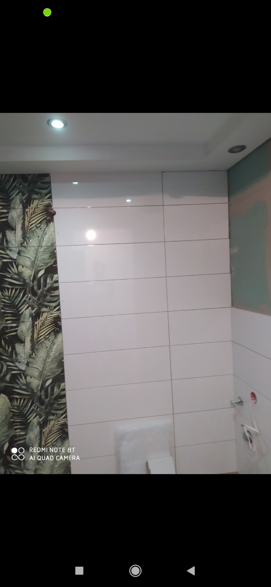 Łazienka w trakcie wykańczania: białe, błyszczące płytki ułożone poziomo na ścianie, fragment ściany z tapetą w tropikalne liście, widoczne niedokończone przyłącza wodne i materiały budowlane...