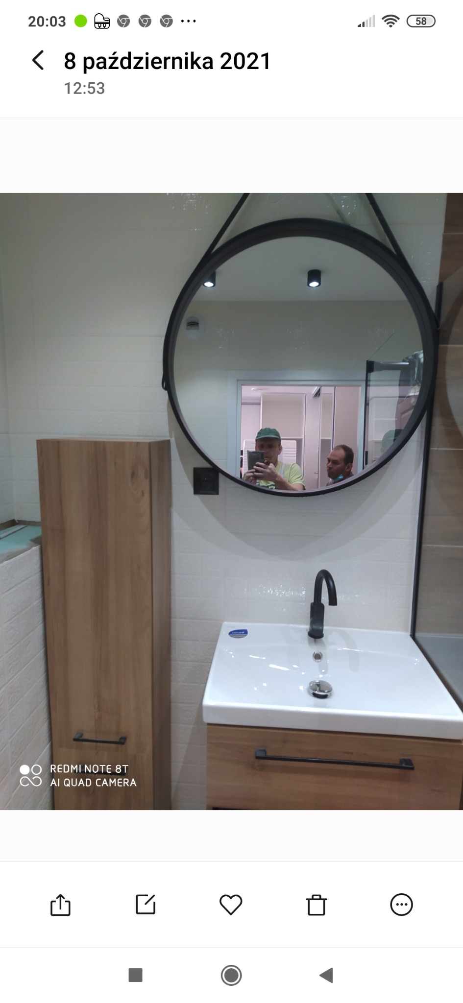 Ukończona łazienka z umywalką na szafce z drewnianym frontem, wiszącą szafką obok i okrągłym lustrem, w którym odbijają się dwie osoby.