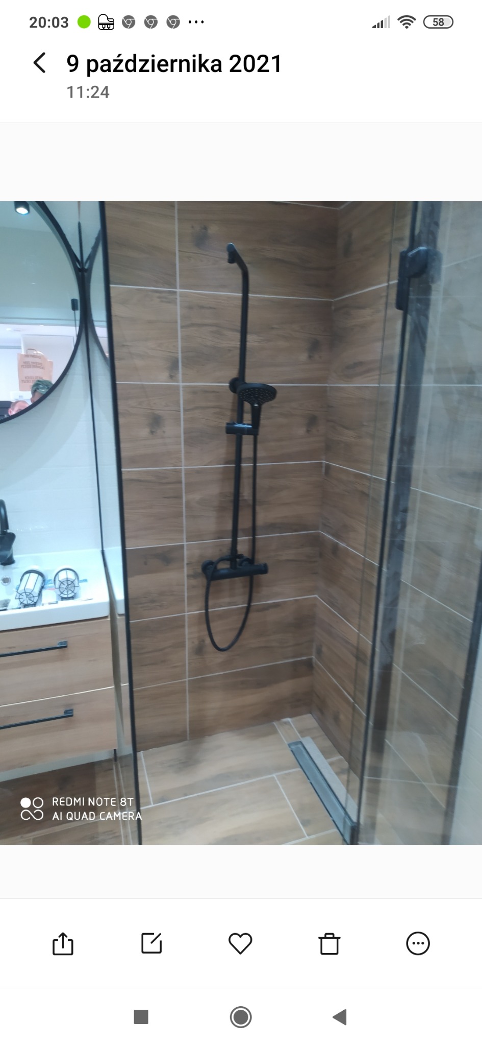 Nowoczesna kabina prysznicowa z czarną armaturą i odpływem liniowym, wykończona płytkami imitującymi drewno, z widoczną umywalką i lustrem w tle.