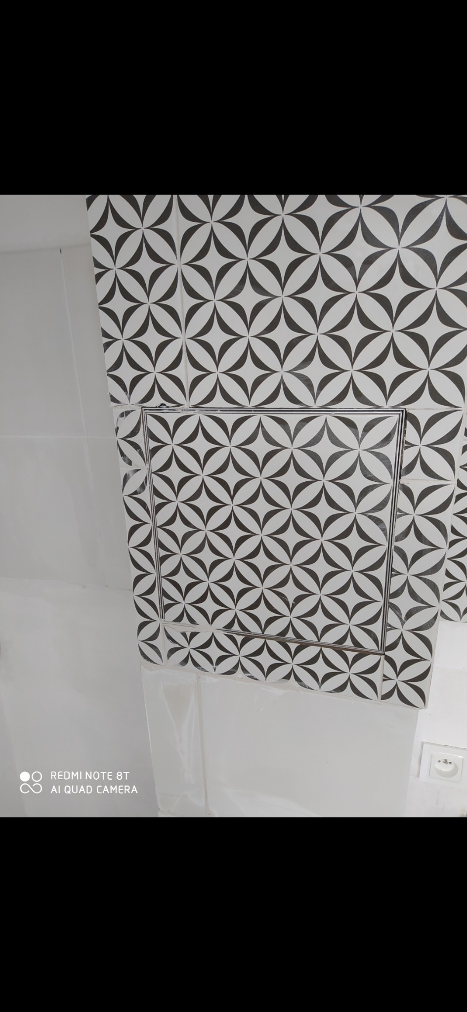 Biała łazienka z czarno-białymi płytkami w geometryczny wzór, widoczny otwór rewizyjny w płytkach.