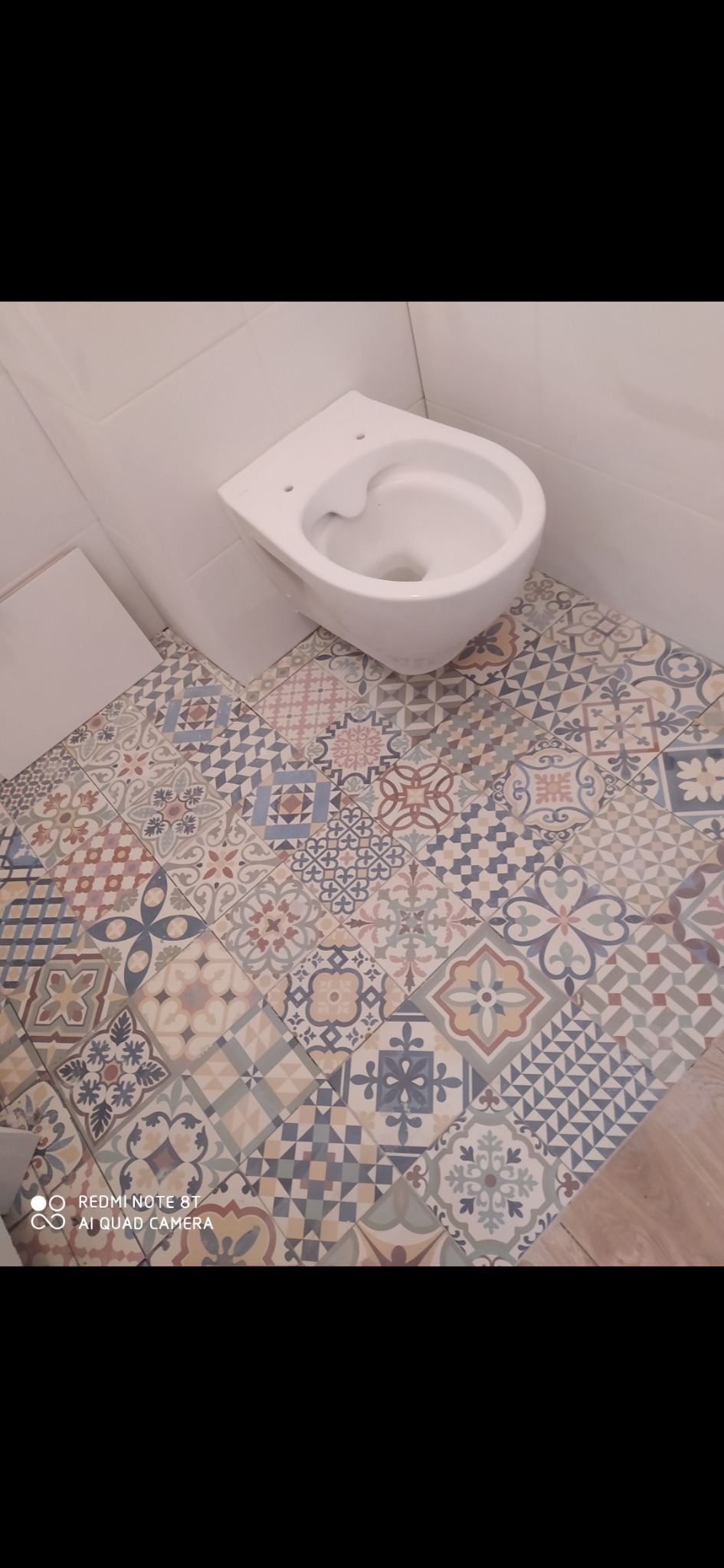 Wisząca muszla klozetowa zamontowana nad nową, wzorzystą podłogą w łazience, widoczne białe płytki na ścianie.