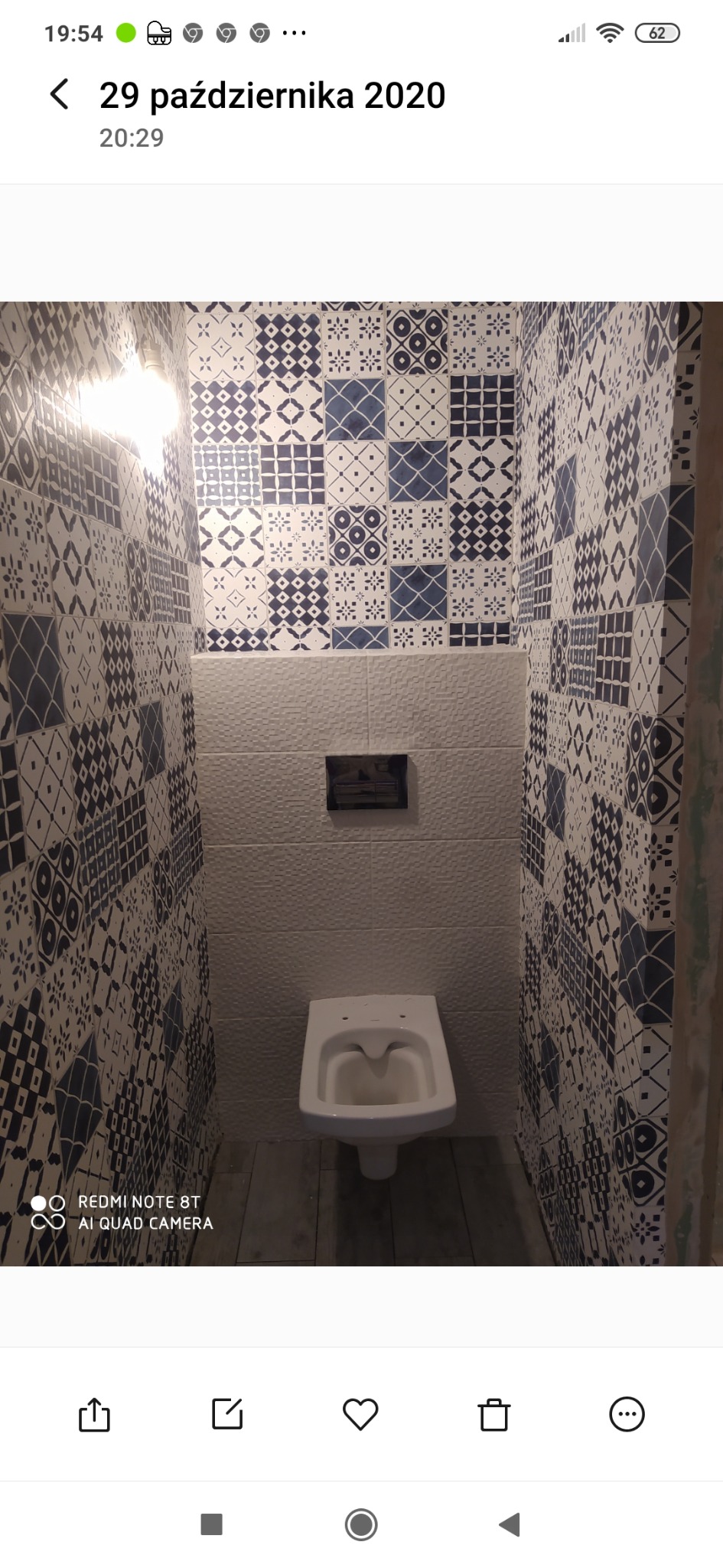 Wnętrze małej toalety z białą miską ustępową wiszącą na ścianie. Ściany wyłożone biało-granatowymi płytkami o wzorach geometrycznych. Widoczne nierówne wykończenie ściany po prawej stronie.