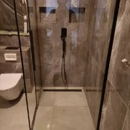 Nowoczesna kabina prysznicowa typu walk-in z czarną armaturą, wykończona płytkami imitującymi marmur, z wnęką w ścianie na kosmetyki i odpływem liniowym w podłodze. Widoczna toaleta w tle.