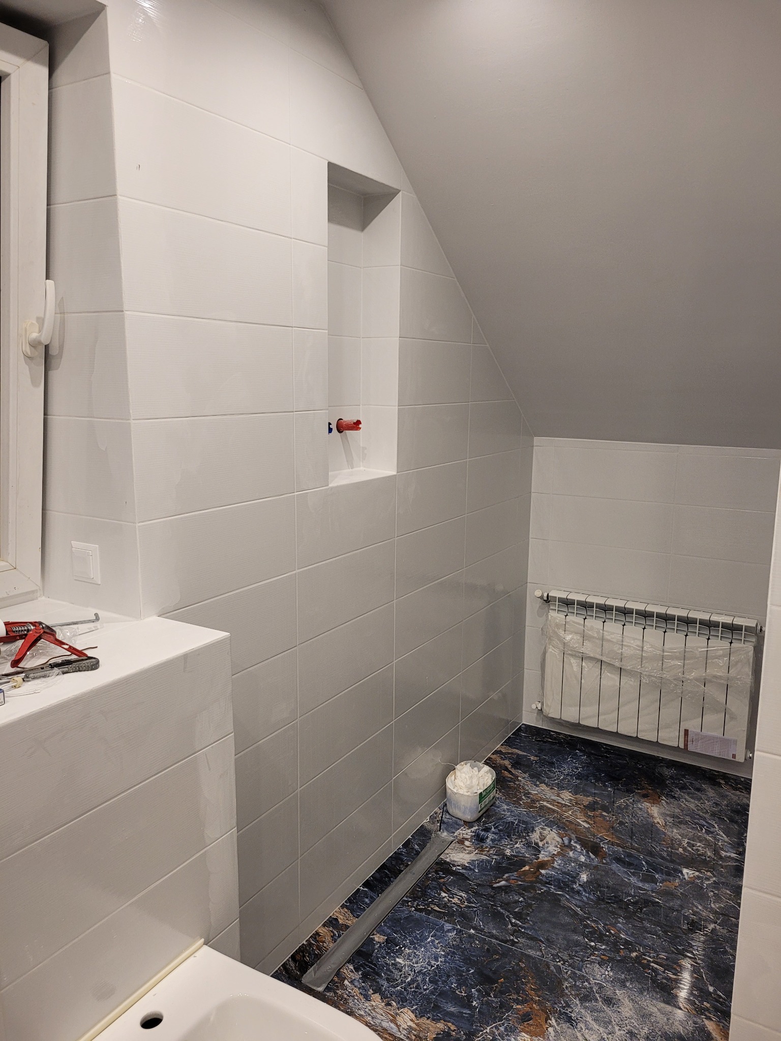 Wykończona łazienka na poddaszu z białą, poziomą glazurą, marmurkową podłogą, grzejnikiem owiniętym folią i narzędziami na parapecie okna.