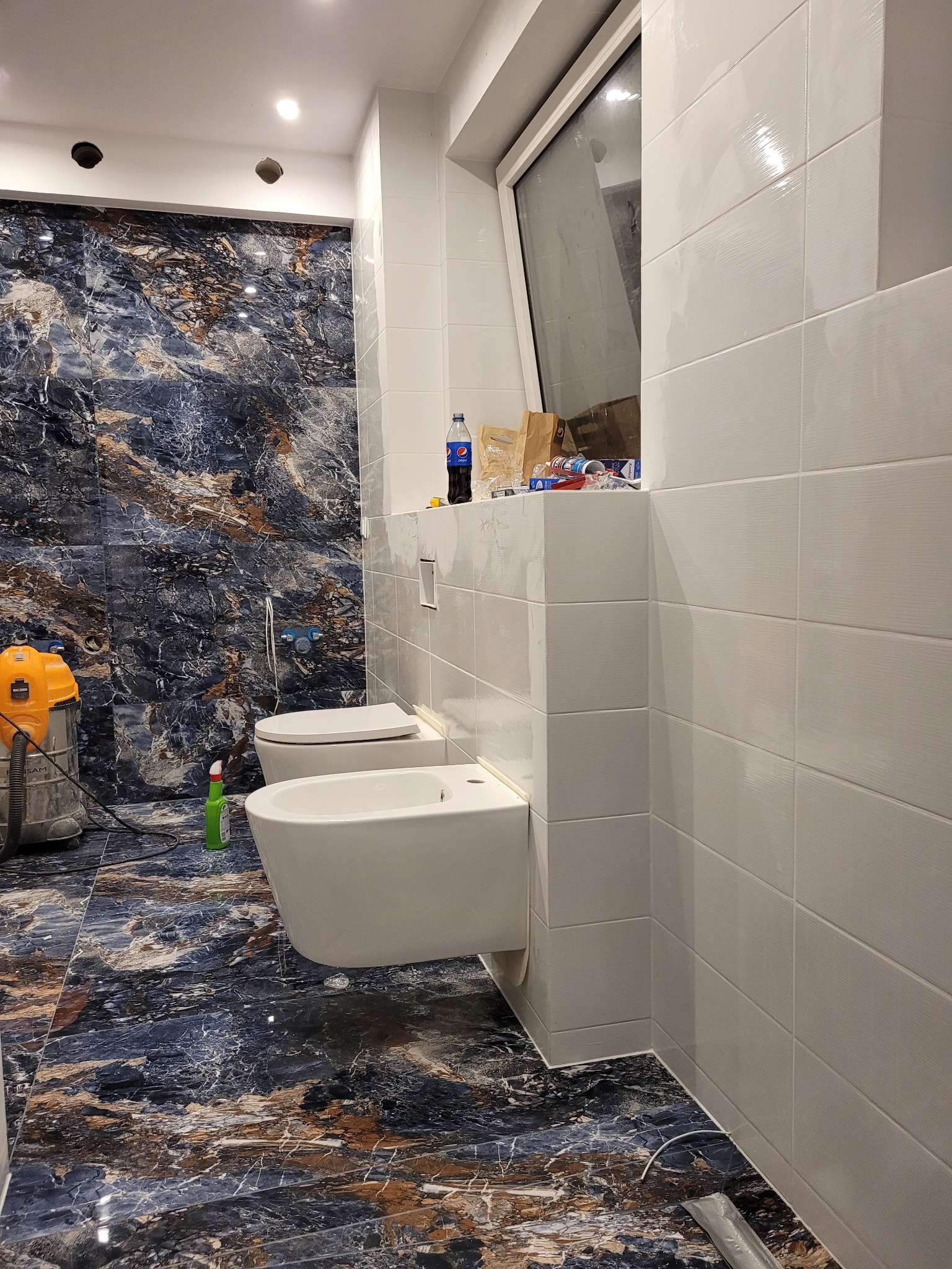 Nowoczesna łazienka w trakcie wykańczania: białe płytki na ścianach, marmurowe płytki podłogowe w odcieniach granatu i brązu, wisząca toaleta i bidet, odkurzacz przemysłowy w rogu, butelka napoju...