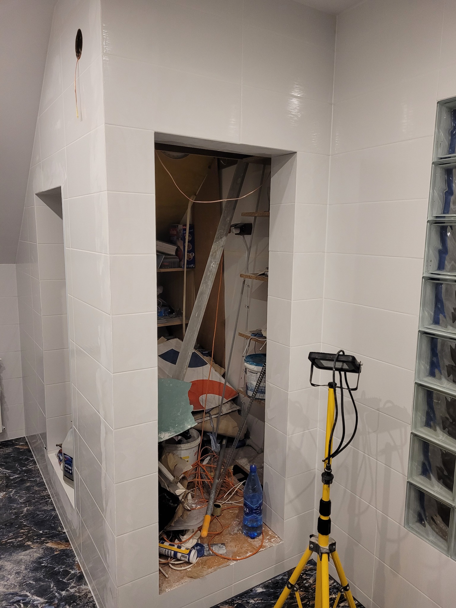 Białe płytki ścienne w trakcie układania, widoczny otwór w ścianie z narzędziami i materiałami budowlanymi w środku, oraz żółty statyw z lampą oświetlającą miejsce pracy.