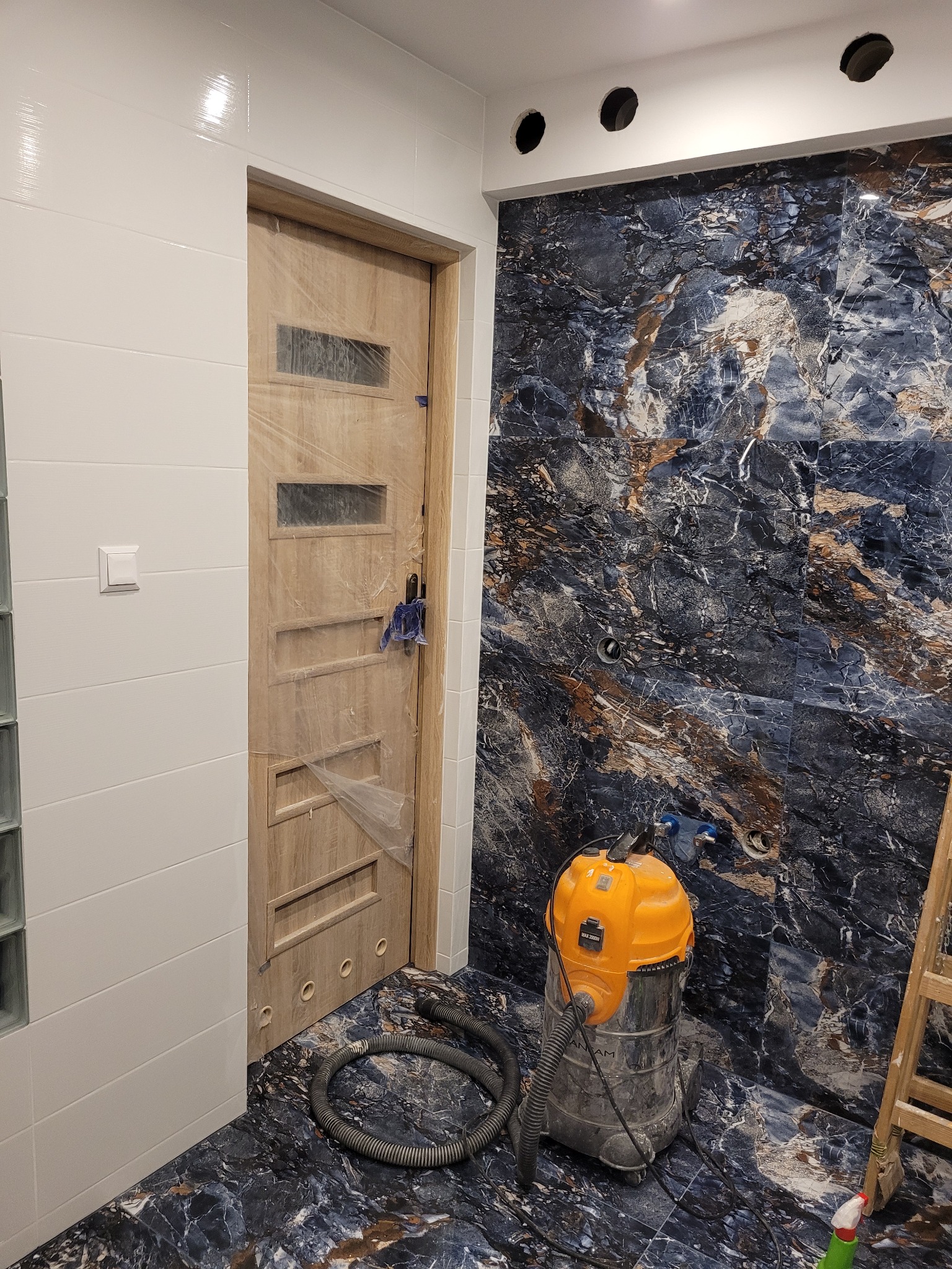 Łazienka w trakcie prac wykończeniowych: położone marmurowe płytki, zabezpieczone folią drzwi, odkurzacz przemysłowy i drabina.