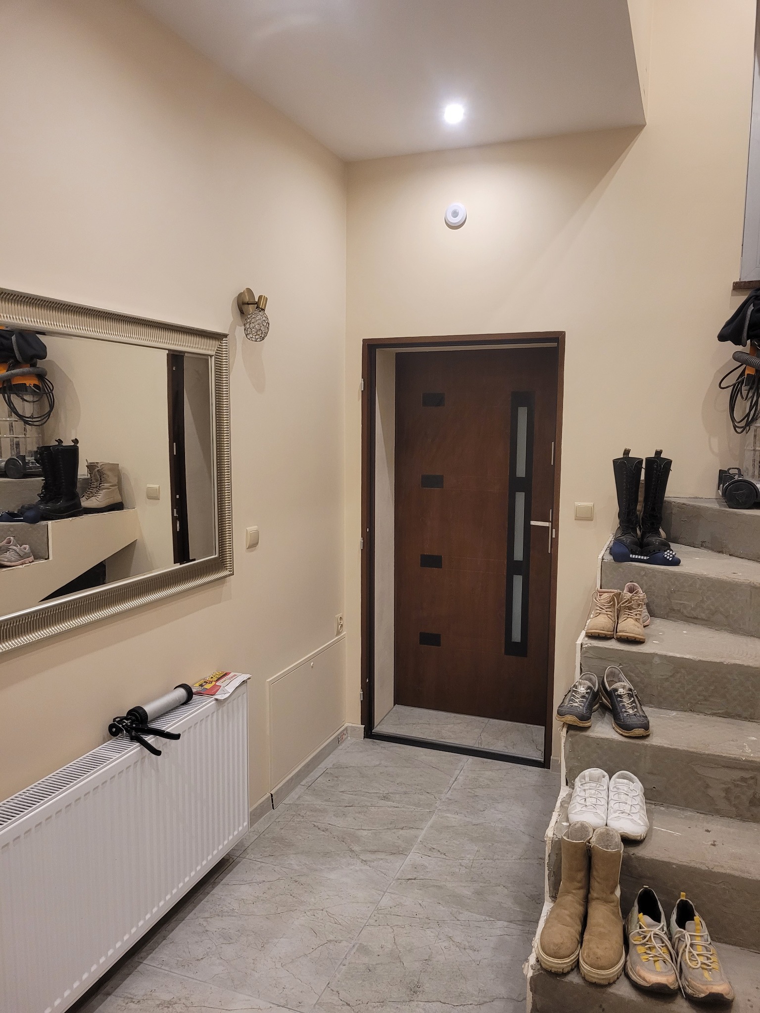 Korytarz z brązowymi drzwiami wejściowymi, grzejnikiem z pistoletem do silikonu, lustrem i schodami, na których leżą buty.