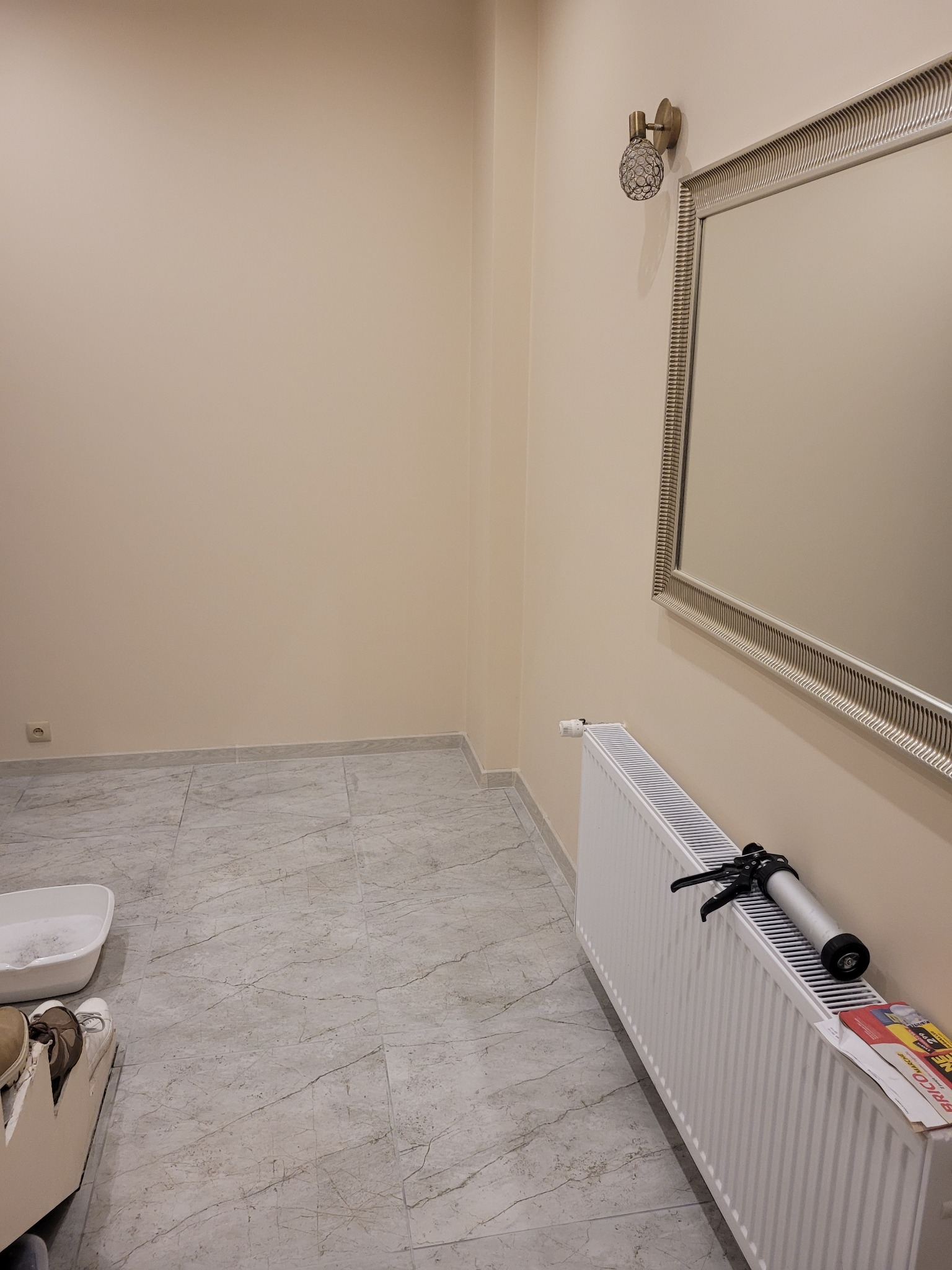 Pusta, jasna ściana z zamontowanym lustrem w ozdobnej ramie i kinkietem, na podłodze marmurowe płytki, widoczny biały grzejnik z pistoletem do silikonu i ulotką reklamową.