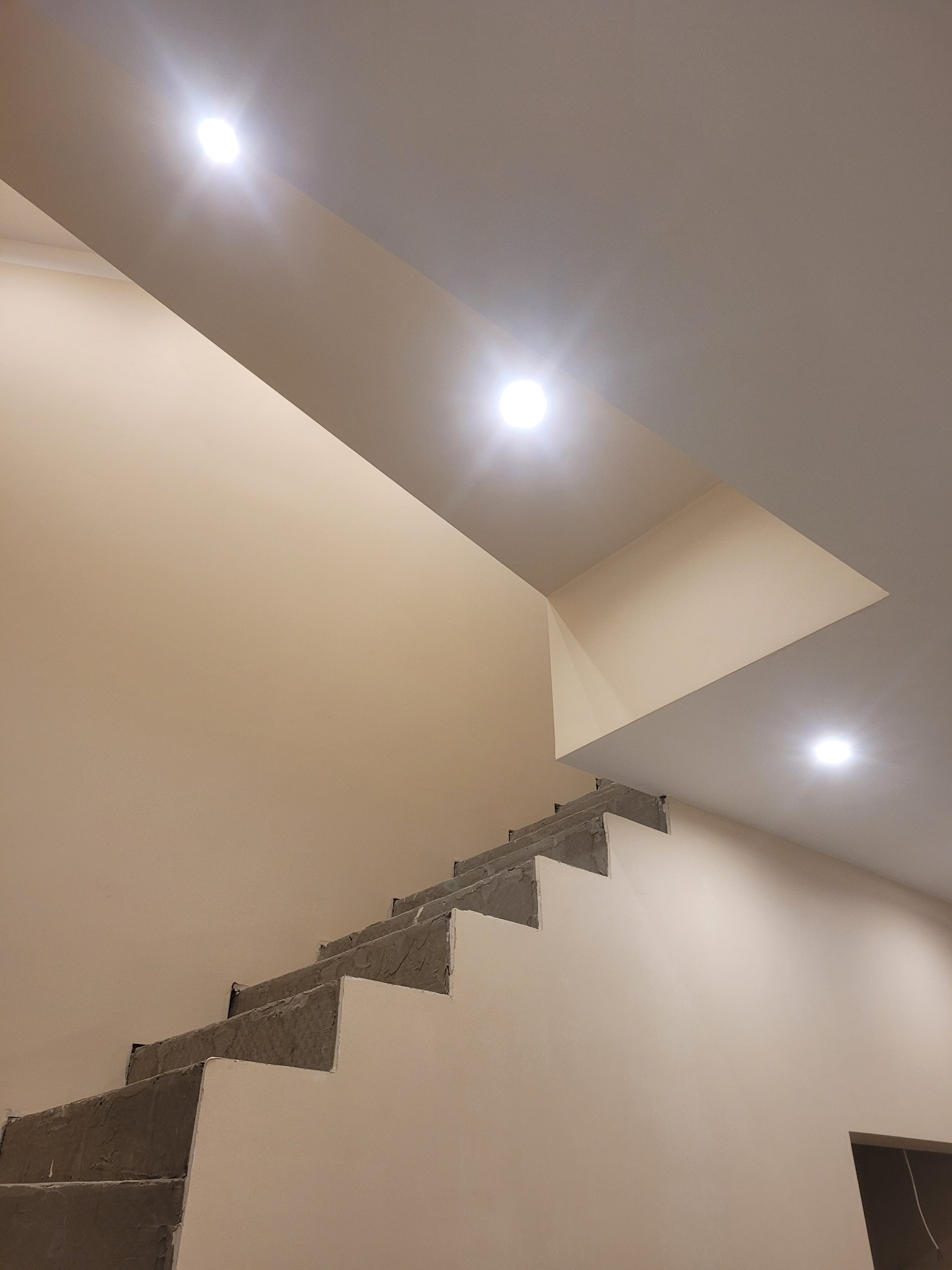 Surowe schody betonowe w trakcie wykańczania, widoczne spodnie stopnie i fragment ściany w jasnym kolorze, oświetlone trzema punktowymi lampami sufitowymi.