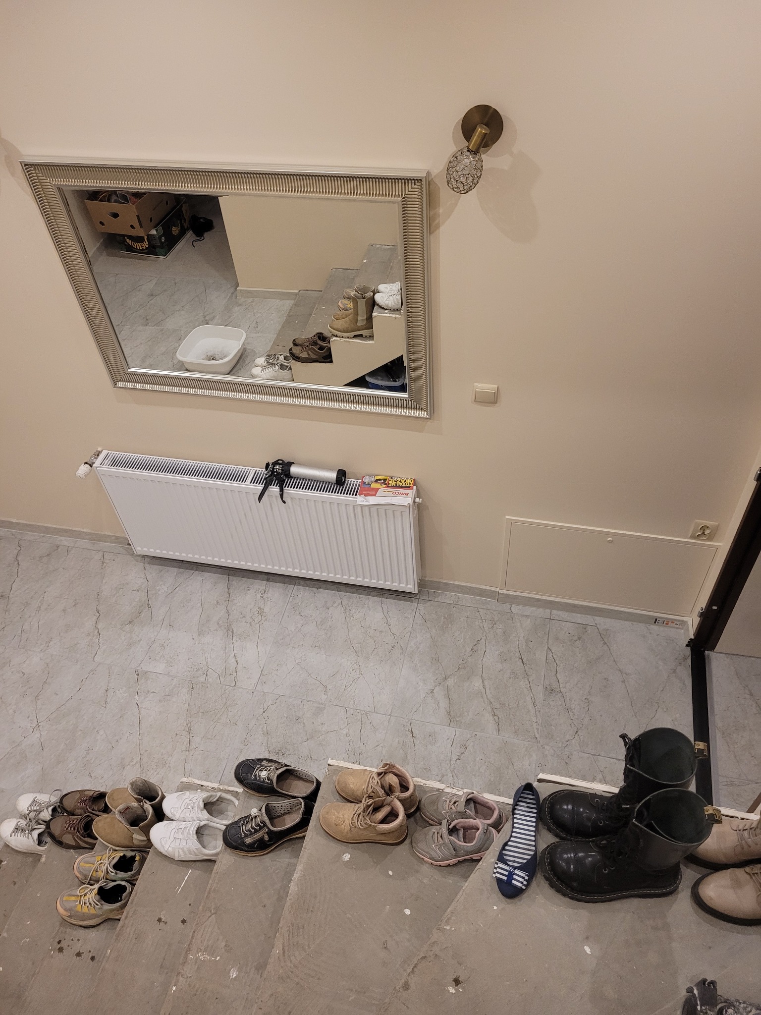 Schody w trakcie remontu z różnym obuwiem na stopniach, grzejnik z narzędziami i lustro na ścianie.