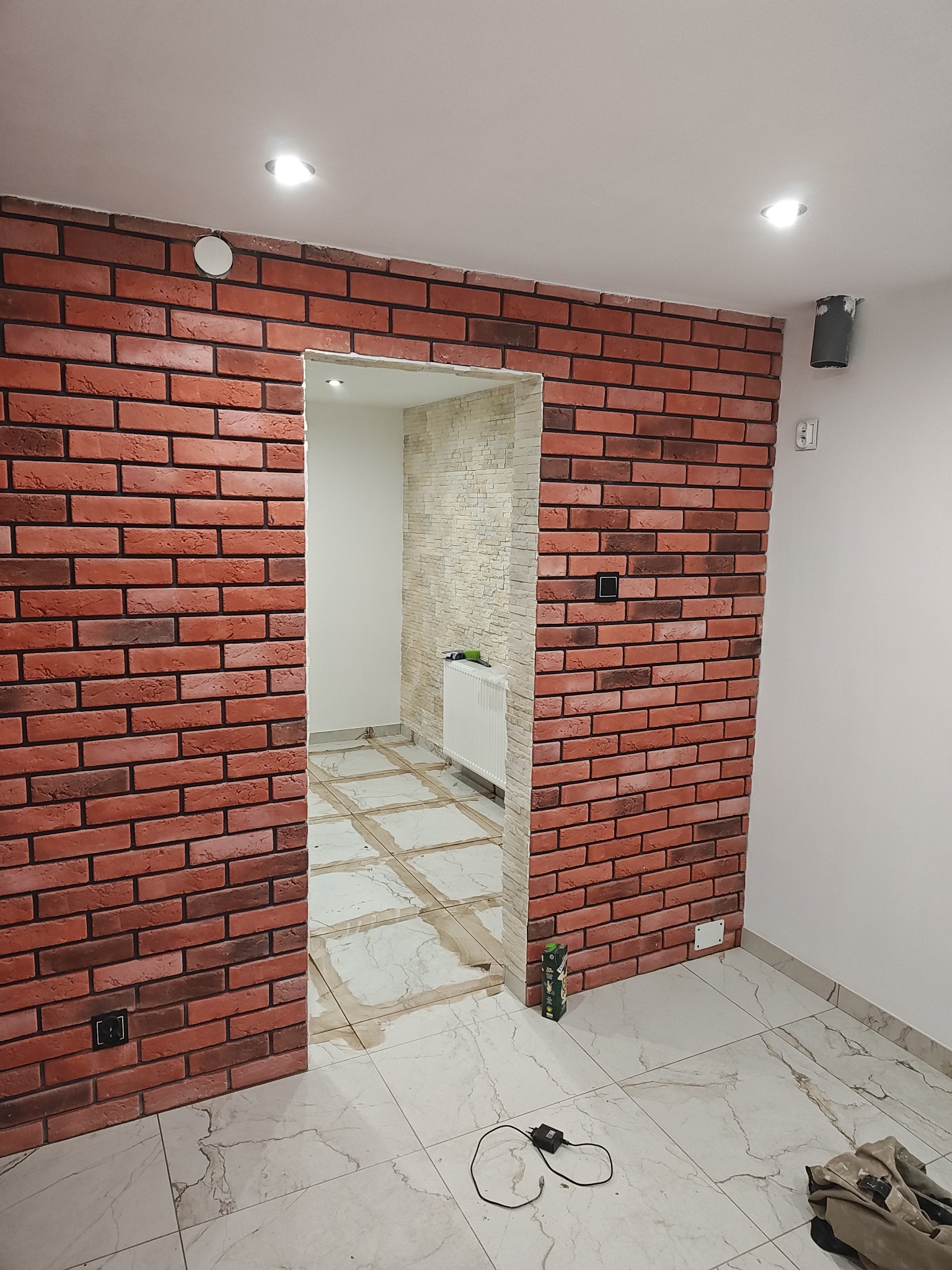 Ściana z czerwonej cegły w trakcie prac wykończeniowych, widoczne przejście do drugiego pomieszczenia z jasną cegłą dekoracyjną, podłoga z marmurowych płytek częściowo zabezpieczona folią.