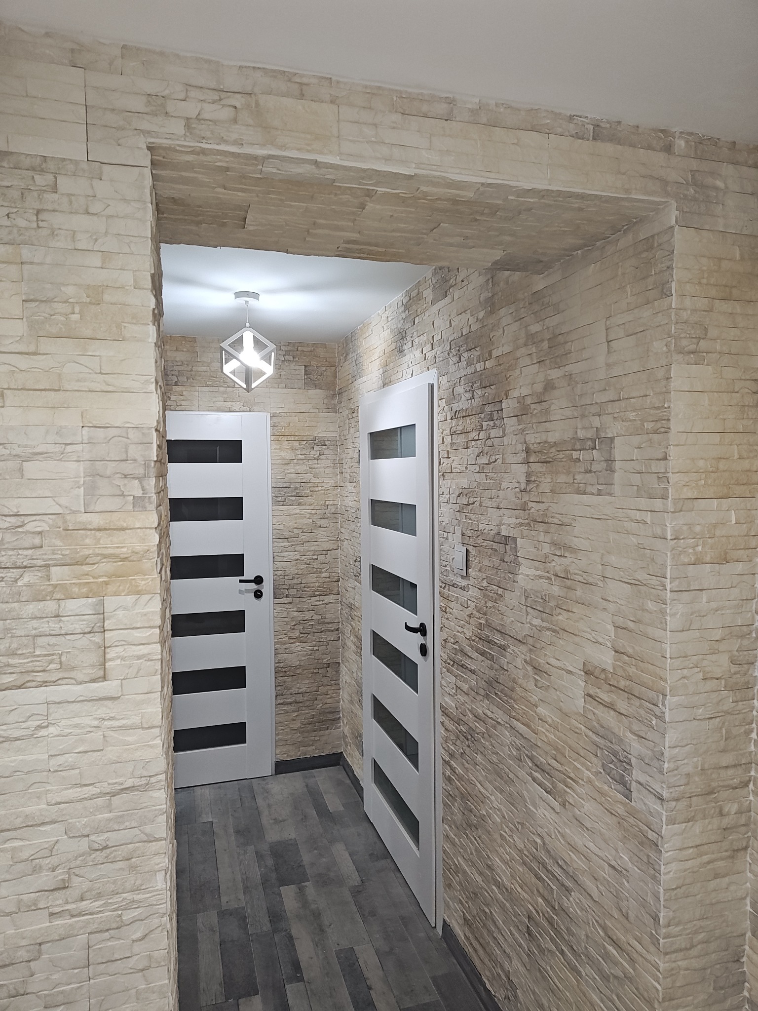 Korytarz z dwiema białymi drzwiami z czarnymi szybami, ściany wyłożone kamieniem dekoracyjnym w odcieniach beżu i szarości, podłoga w panelach imitujących drewno, nowoczesna lampa sufitowa.