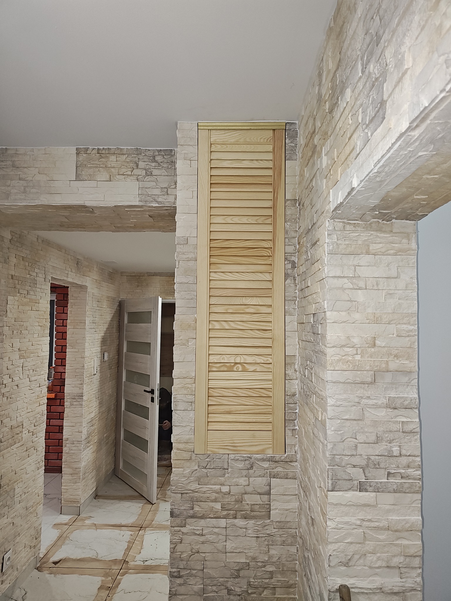 Wykończone wnętrze z dekoracyjną ścianą z kamienia imitującego cegłę, drewnianymi żaluzjowymi drzwiczkami rewizyjnymi i otwartymi drzwiami z przeszkleniami w tle.