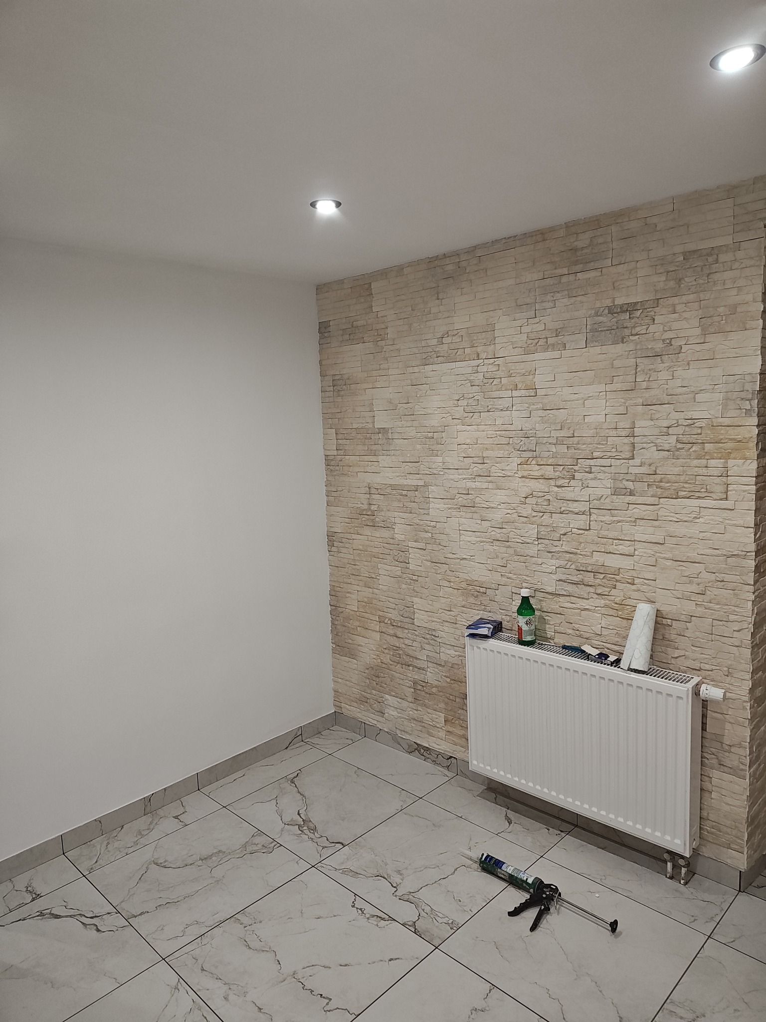 Pokój po częściowym remoncie: ściana z dekoracyjnej cegły, biała ściana, marmurowe płytki podłogowe, grzejnik z akcesoriami i pistolet do silikonu na podłodze.