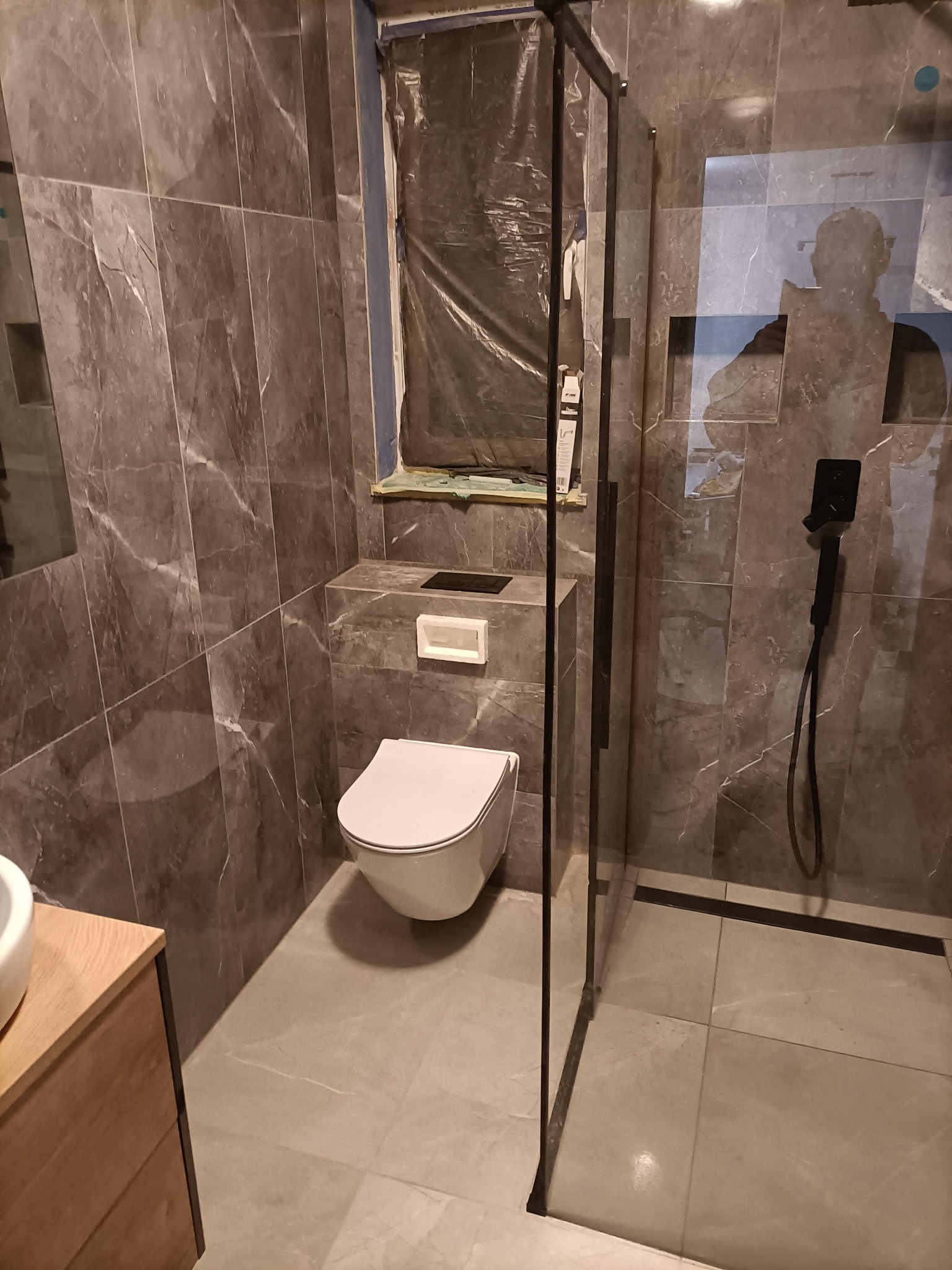 Wykończona łazienka z marmurowymi płytkami, kabiną prysznicową typu walk-in z czarnymi profilami, podwieszaną toaletą i oknem zabezpieczonym folią malarską podczas remontu.