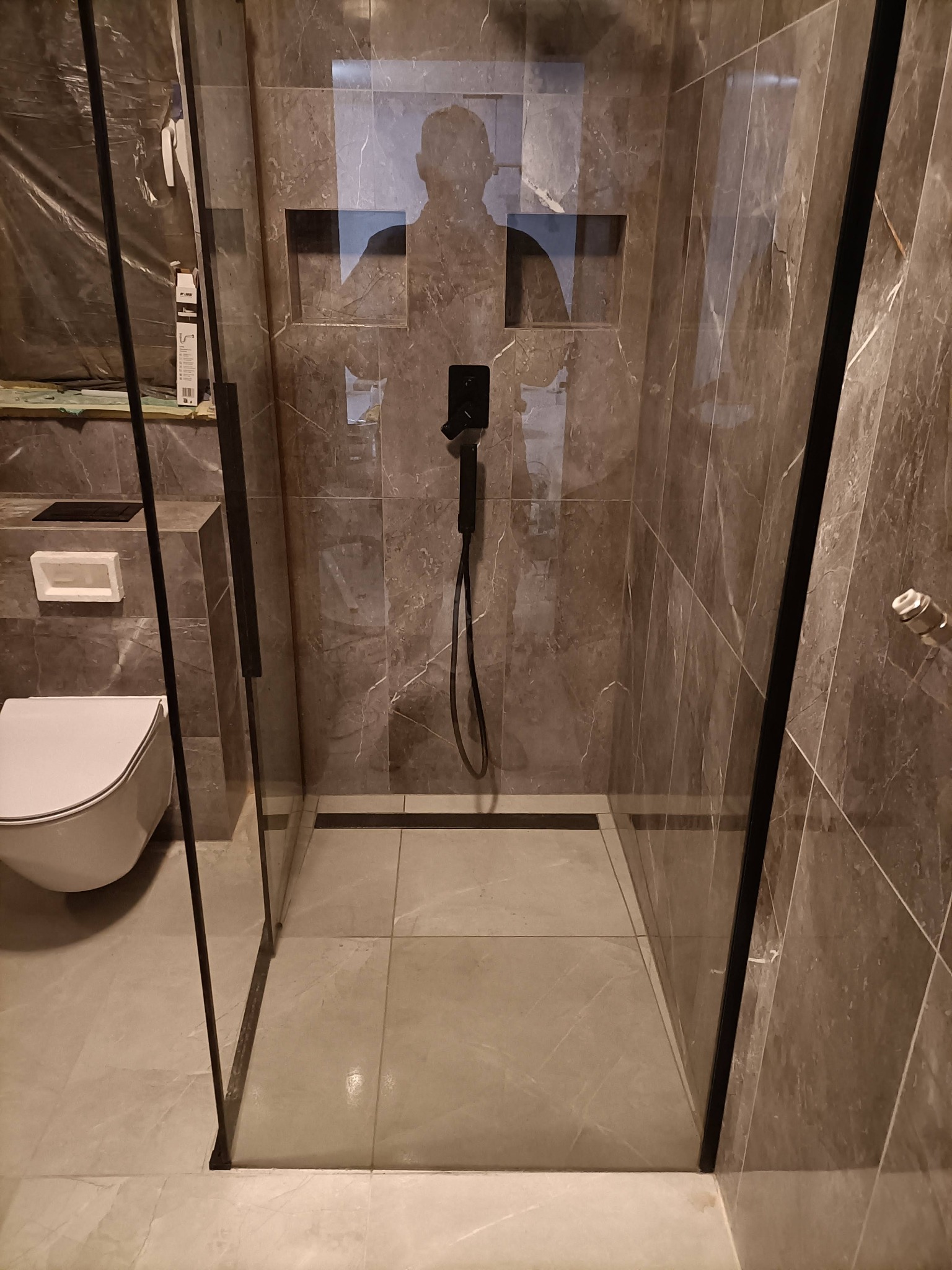 Nowoczesna kabina prysznicowa typu walk-in z czarną armaturą, wykończona płytkami imitującymi marmur, z wnęką w ścianie na kosmetyki i odpływem liniowym w podłodze. Widoczna toaleta w tle.