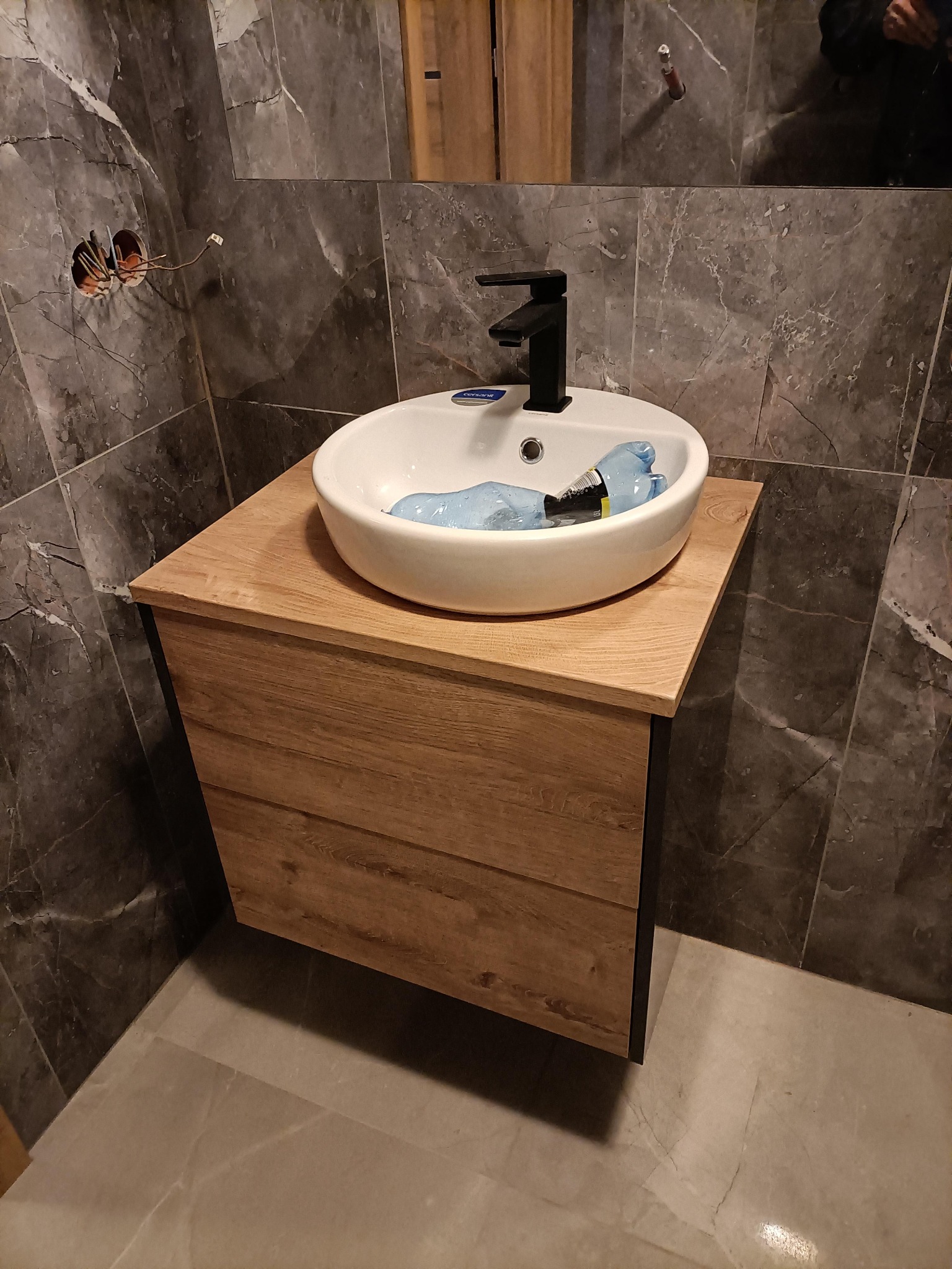 Nowoczesna łazienka z okrągłą umywalką nablatową, czarną armaturą i szafką z drewnopodobnym frontem. Na ścianie widoczne marmurowe płytki i niezamaskowane kable elektryczne.