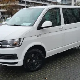 Biały Volkswagen Transporter z czarnymi felgami parkuje na mokrym, brukowanym placu, widoczne drzewa w tle.