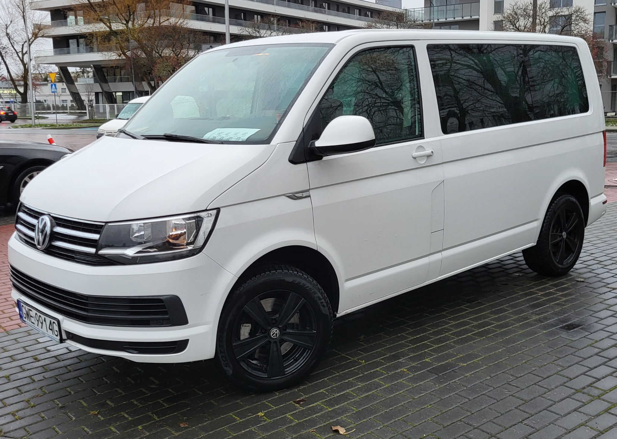 Biały Volkswagen Transporter z czarnymi felgami parkuje na mokrym, brukowanym placu, widoczne drzewa w tle.
