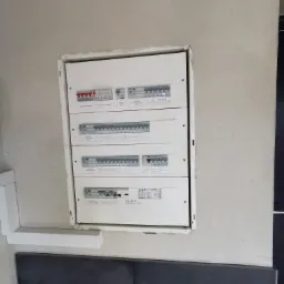 Zamontowana, biała skrzynka rozdzielcza z bezpiecznikami, widoczna na tle jasnej ściany w nowym budynku, z fragmentem ciemnej płytki poniżej.