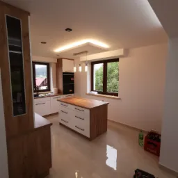 Nowoczesna kuchnia z wyspą, drewnianymi akcentami i oświetleniem LED, widoczne narzędzia i napoje sugerują trwające prace wykończeniowe.