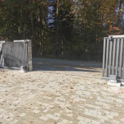 Automatyczna brama przesuwna z szarych paneli, częściowo otwarta, zamontowana na posesji z kostki brukowej pokrytej opadłymi liśćmi, widoczny napęd bramy i surowy murek z bloczków betonowych.