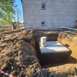 Betonowy zbiornik z kominkiem wentylacyjnym w wykopie zabezpieczonym taśmą ostrzegawczą, w tle budynek mieszkalny i koparka.