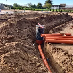 Instalacja rur kanalizacyjnych w wykopie z czarną studzienką kanalizacyjną z czerwoną poziomicą na pokrywie, widok z ziemi na wykopane rowy i fundamenty budynku w tle.