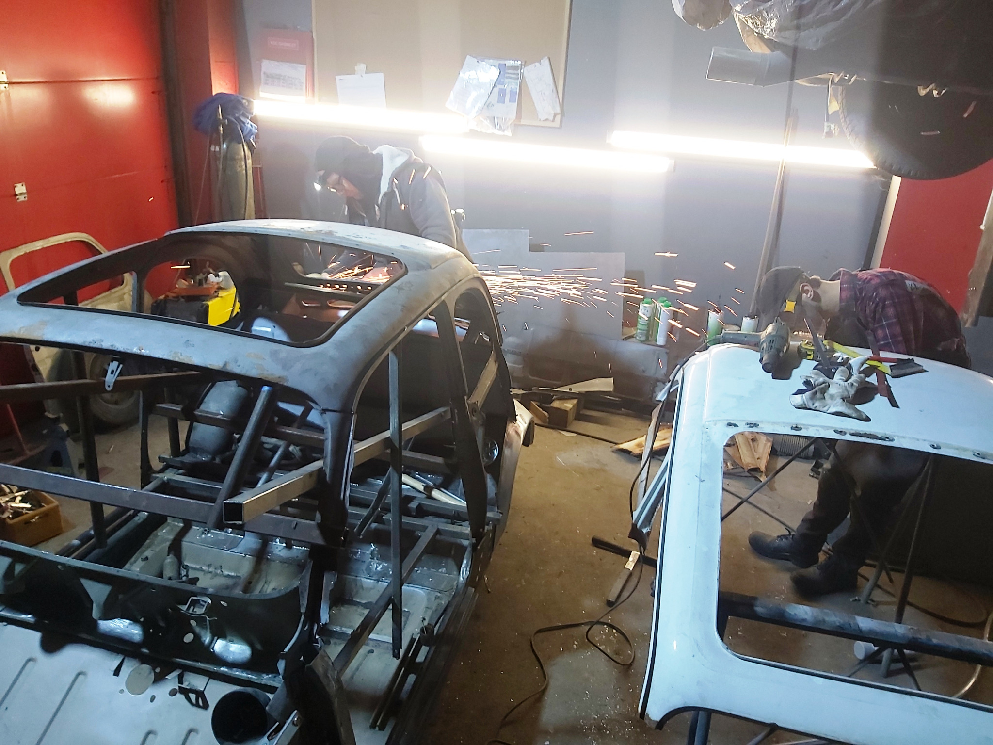 Warsztatowa scena z dwoma mechanikami pracującymi nad karoserią samochodu: jeden spawa, iskrząc, szkielet pojazdu, drugi obrabia dach, w tle widoczne narzędzia i butle.