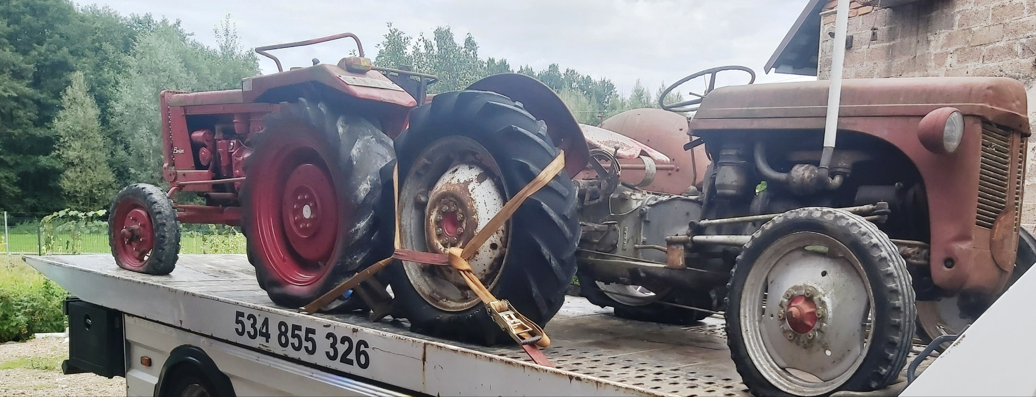 Dwa stare, czerwone traktory przewożone na lawecie, jeden z uszkodzonym kołem zabezpieczonym pasami transportowymi.