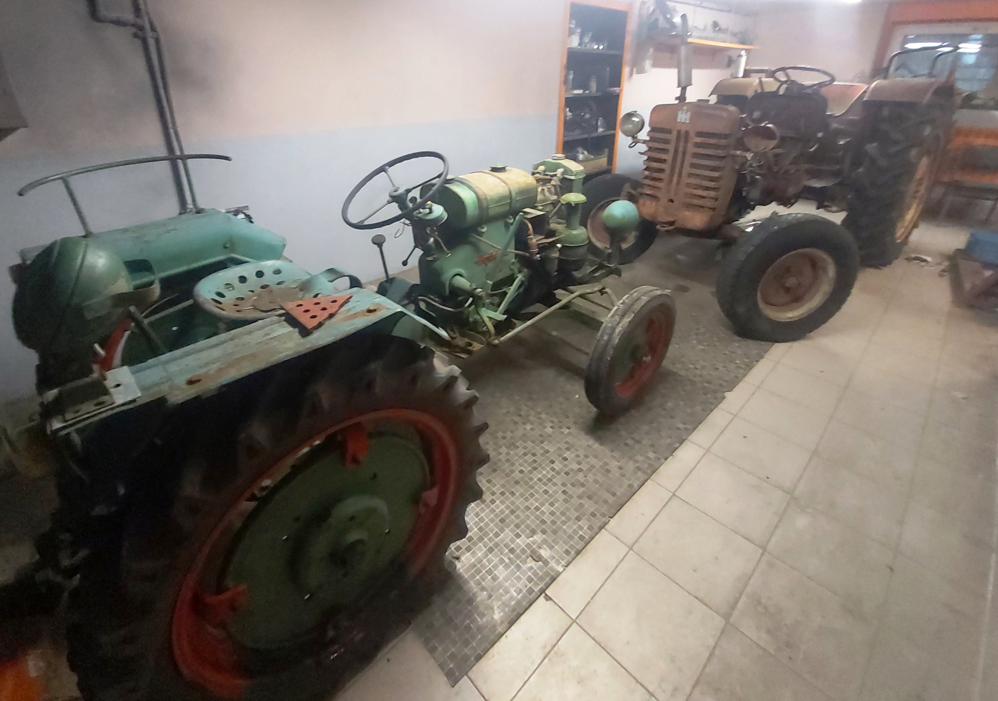 Dwa stare, zielone traktory oraz jeden rdzawy traktor w garażu z podłogą wyłożoną kafelkami.