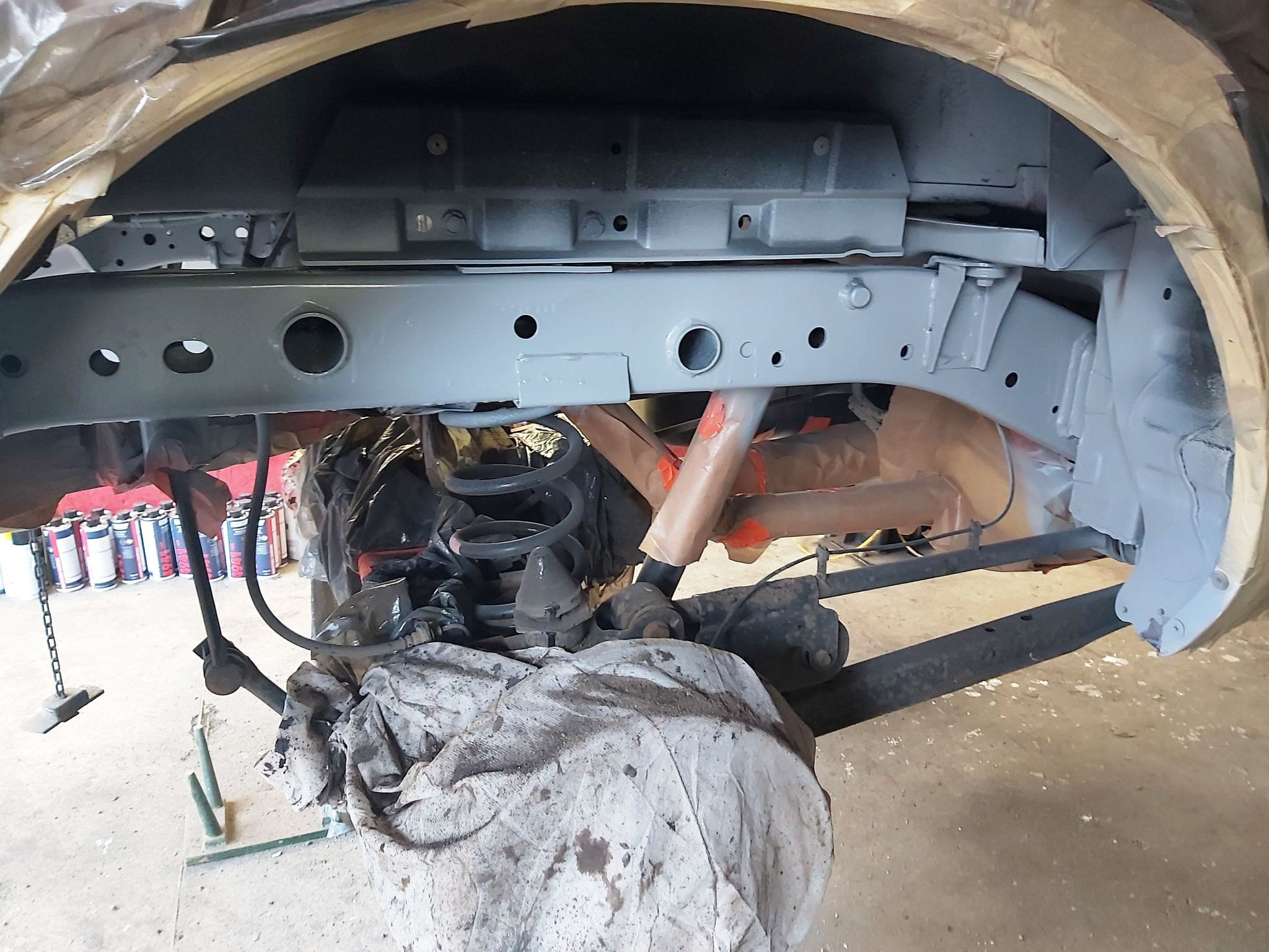 Podwozie samochodu po zabezpieczeniu antykorozyjnym, widoczne elementy zawieszenia i blachy pokryte szarym podkładem, przygotowane do dalszych prac lakierniczych, z boku widoczne puszki z preparatami.