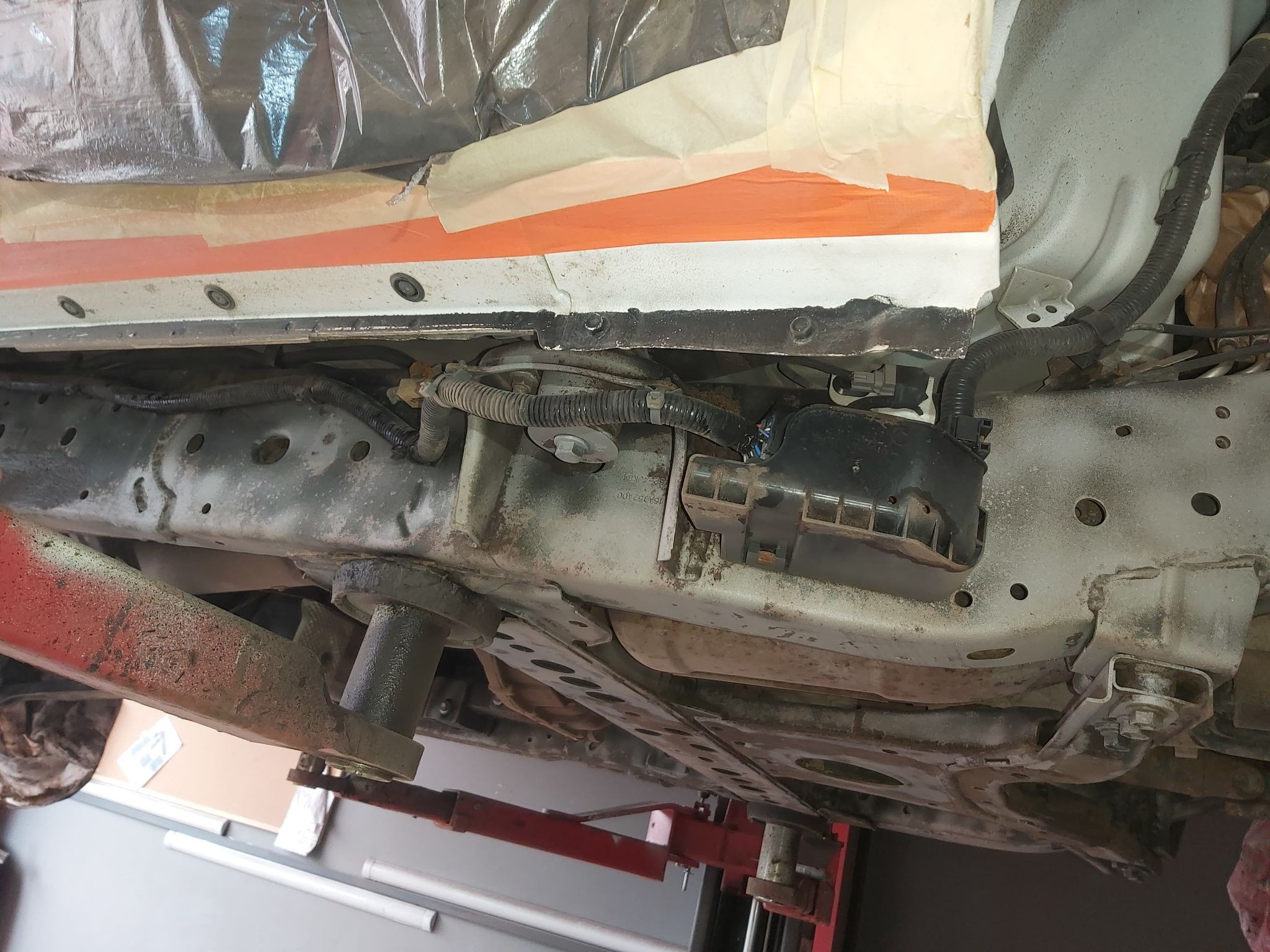 Spód samochodu na podnośniku, widoczne elementy zawieszenia, przewody i zabezpieczenia antykorozyjne w trakcie prac, fragment karoserii oklejony taśmą malarską.